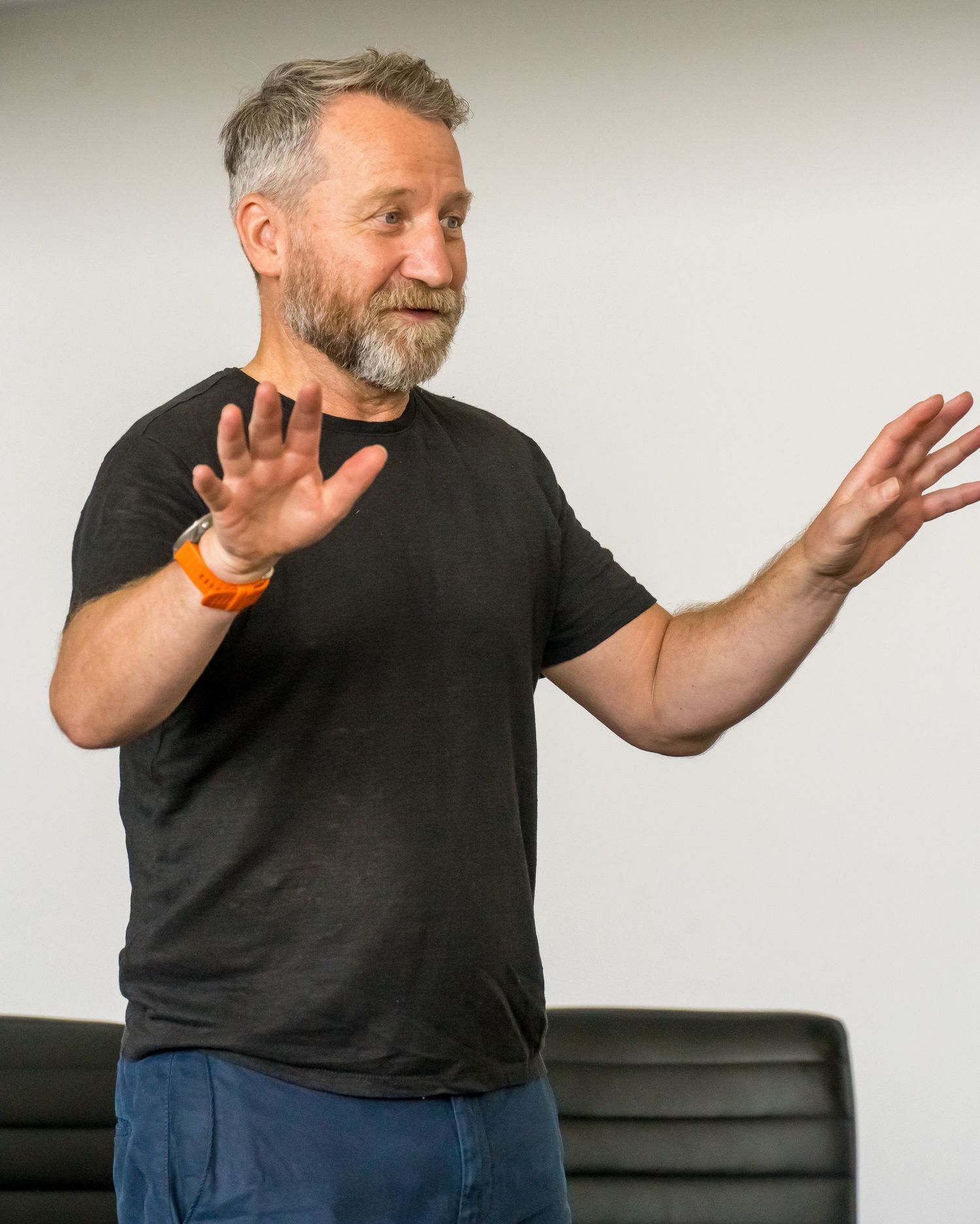 Bearded man with gray hair in a black t-shirt and orange wristband gestures with both hands while speaking in front of a black chair.