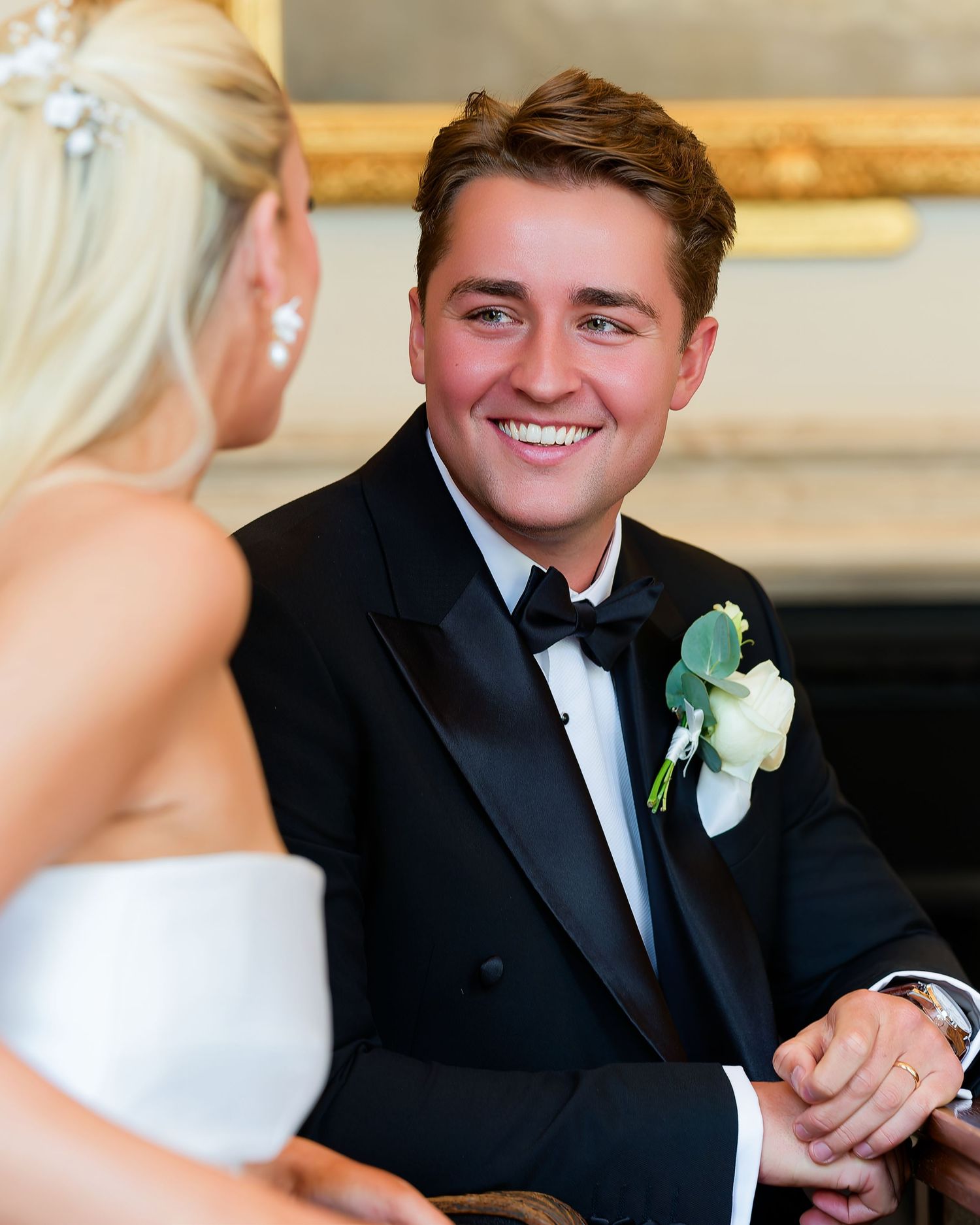 Smiling groom in black tuxedo holding bride's hands during wedding ceremony indoors.