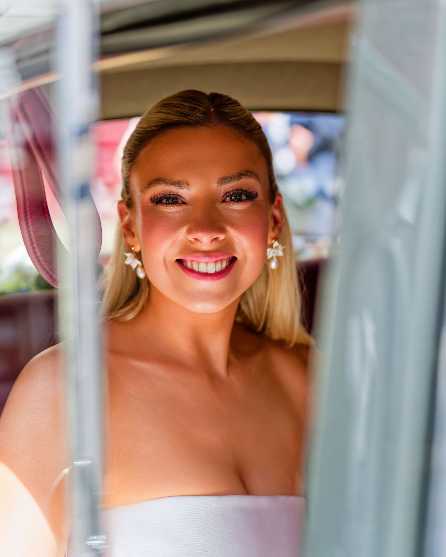Beautiful blonde bride smiles radiantly through vintage car window wearing pearl earrings on wedding day.