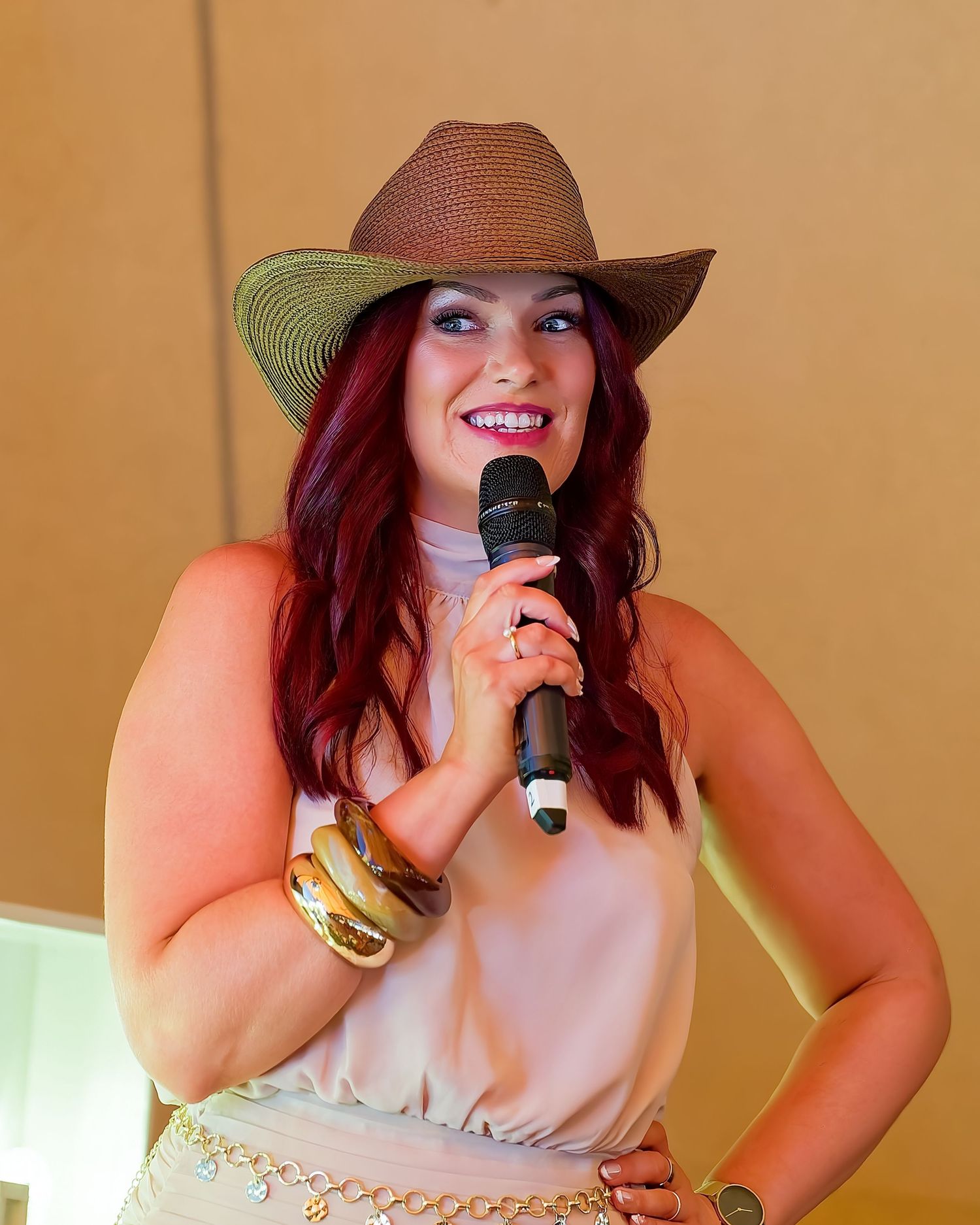 Smiling woman with long auburn hair wears a straw cowboy hat and cream sleeveless top, holding a microphone and stacked gold bangles.