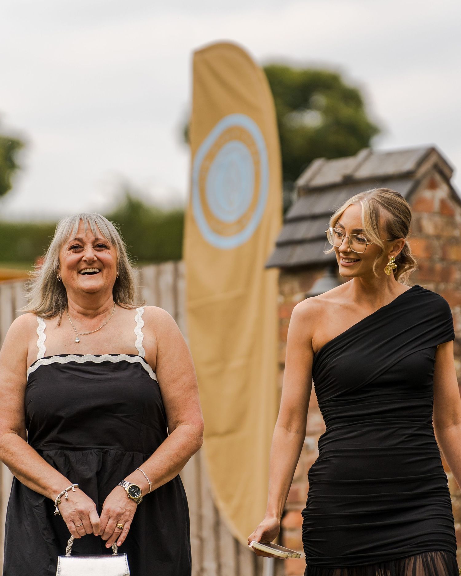 Two smiling women in black dresses stand outdoors at a rustic event, with a tall golden banner and brick building behind them.