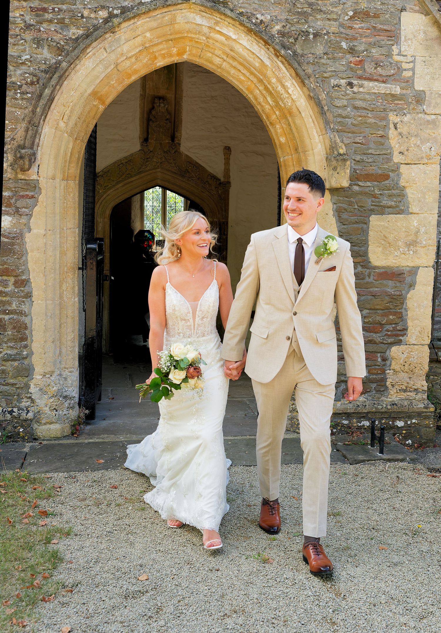 A smiling bride and groom hold hands walking out of a stone church arch, she in white gown, he in tan suit.