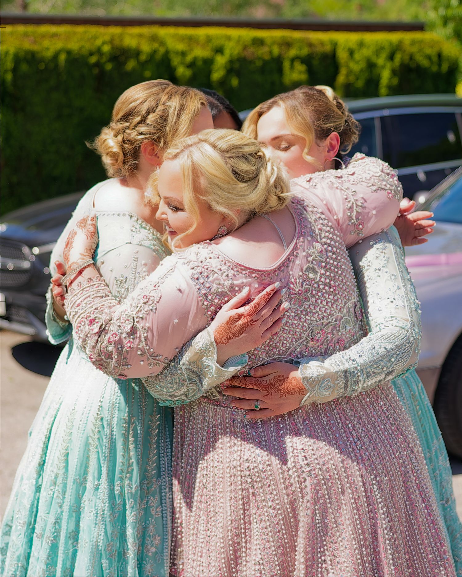 Four women in elegant beaded pastel gowns share a heartfelt group hug outdoors at a formal event.