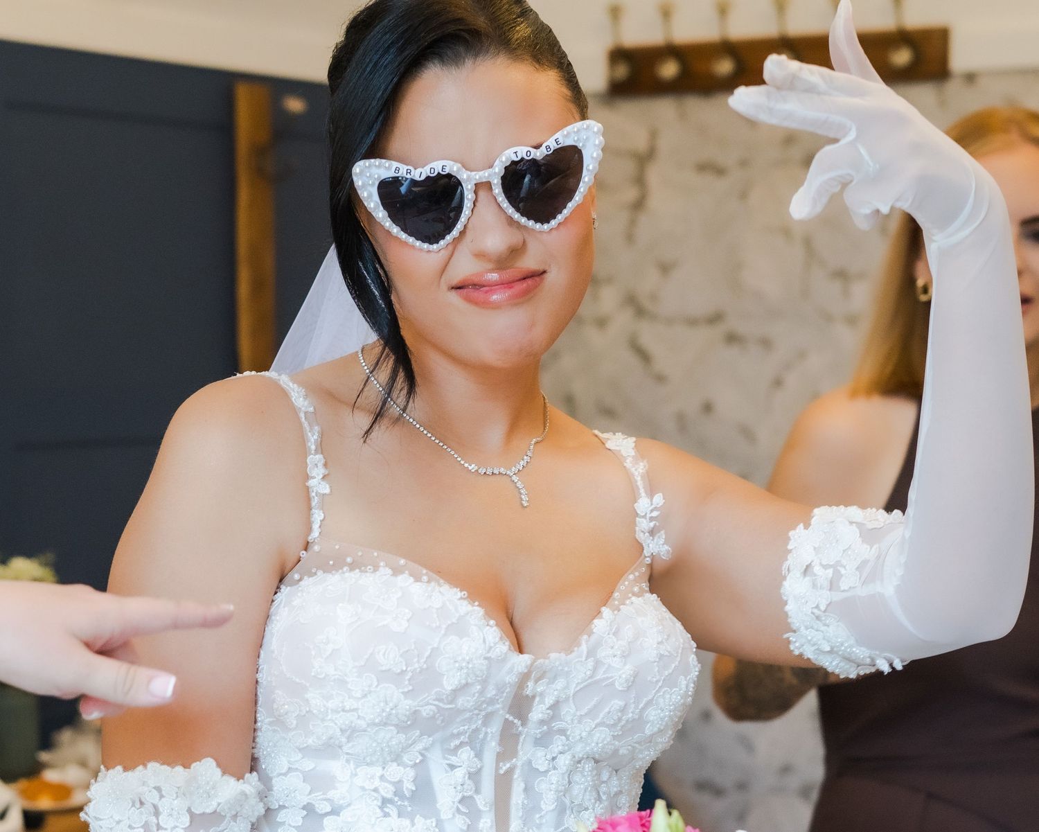 Bride in white lace gown, heart-shaped sunglasses, veil, and white gloves posing playfully at wedding venue.