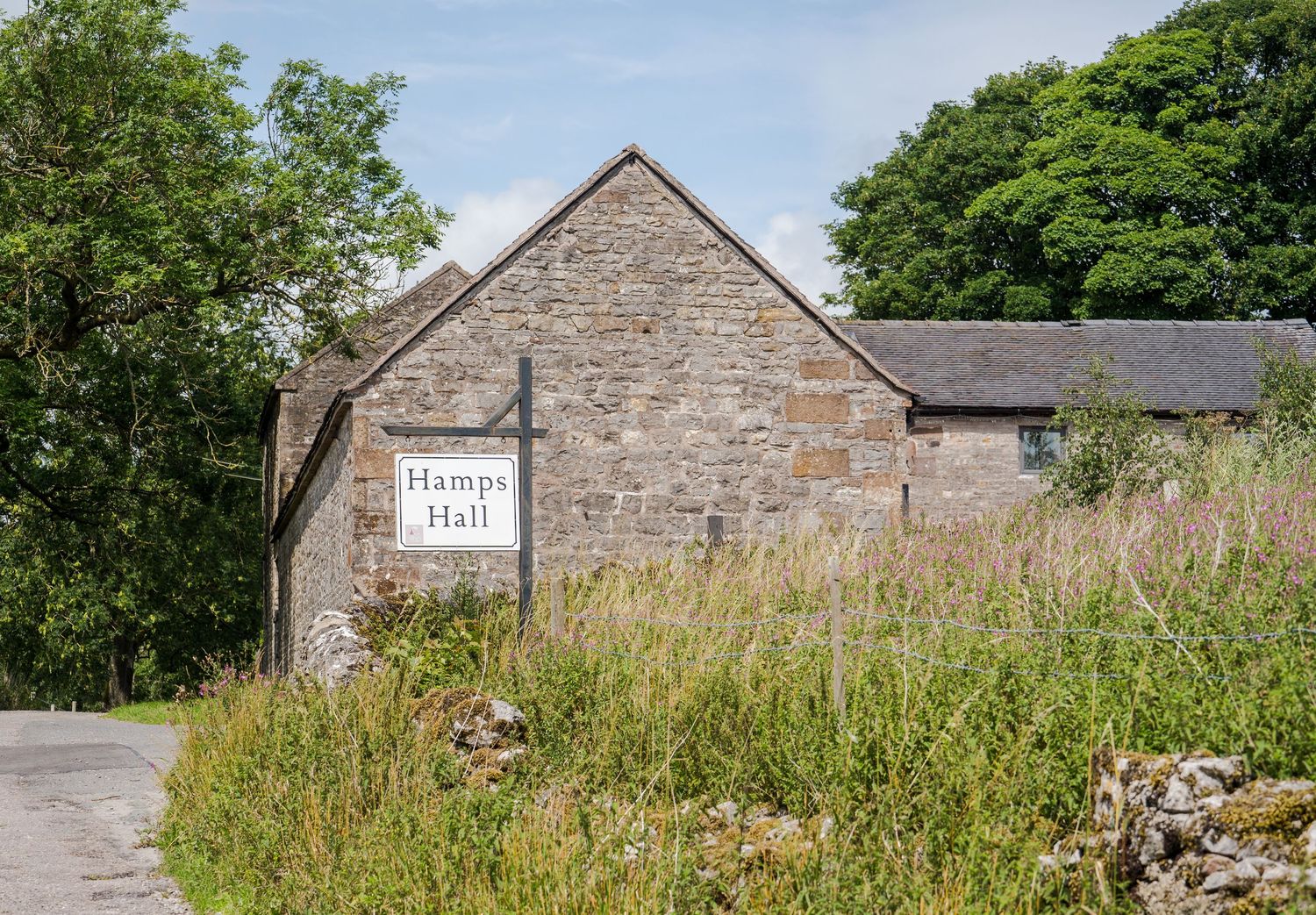A sign reading 'Hamps Hall' mounted on a black post beside a stone wall and country path.