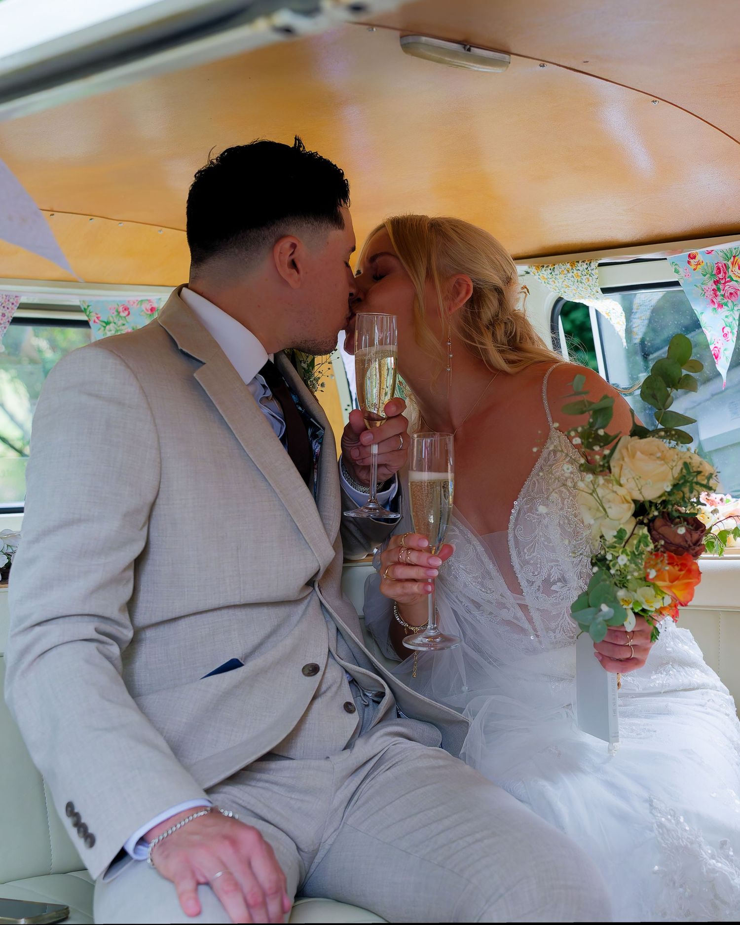 Bride and groom kissing while holding champagne flutes inside a vintage VW campervan on their wedding day.