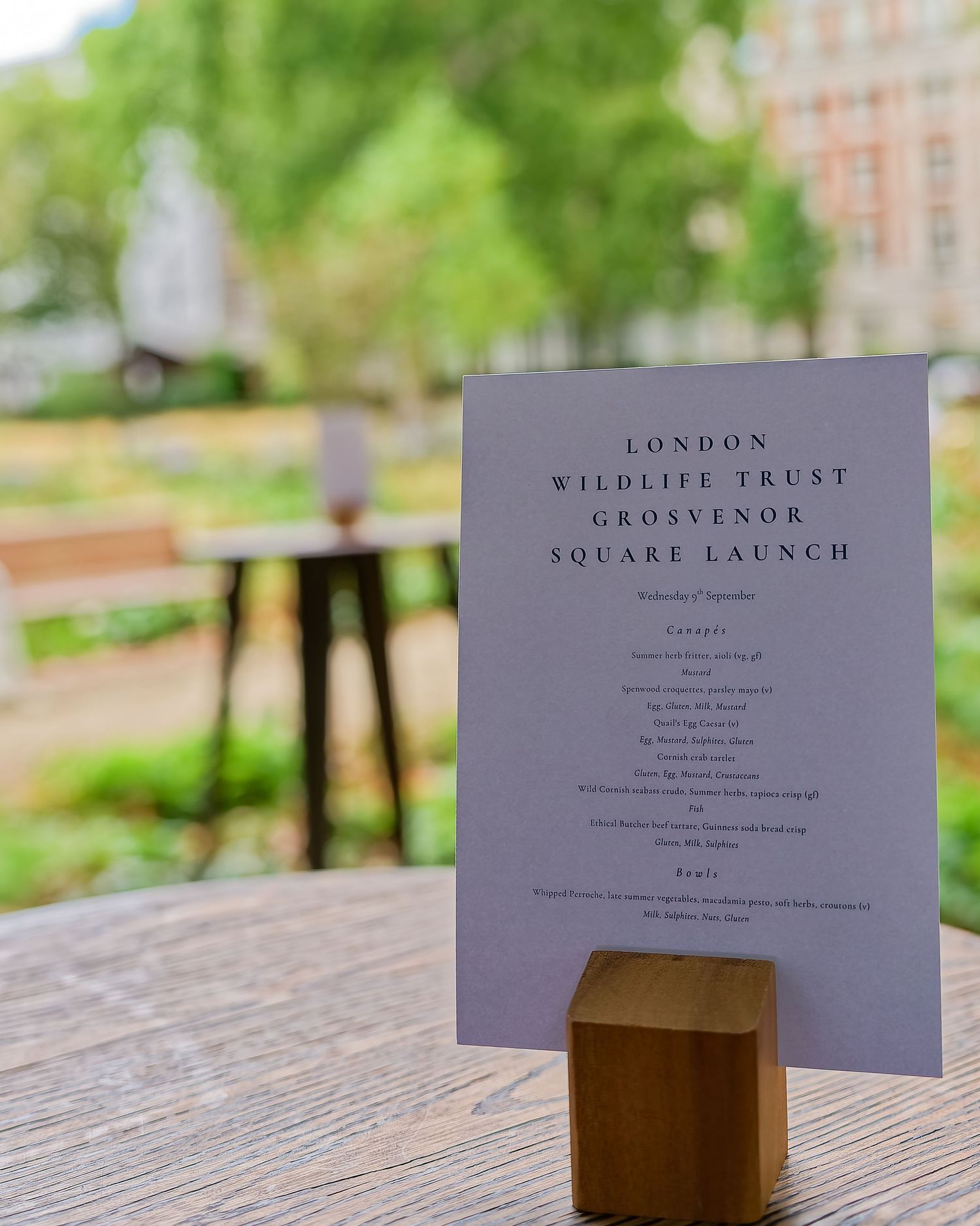 Printed menu for the London Wildlife Trust Grosvenor Square launch in a wooden holder on an outdoor table with a blurred park behind.