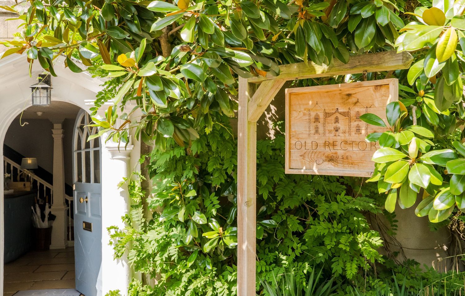 Lush green garden entrance with wooden archway, magnolia trees, and a decorative sign beside a charming white arched doorway.