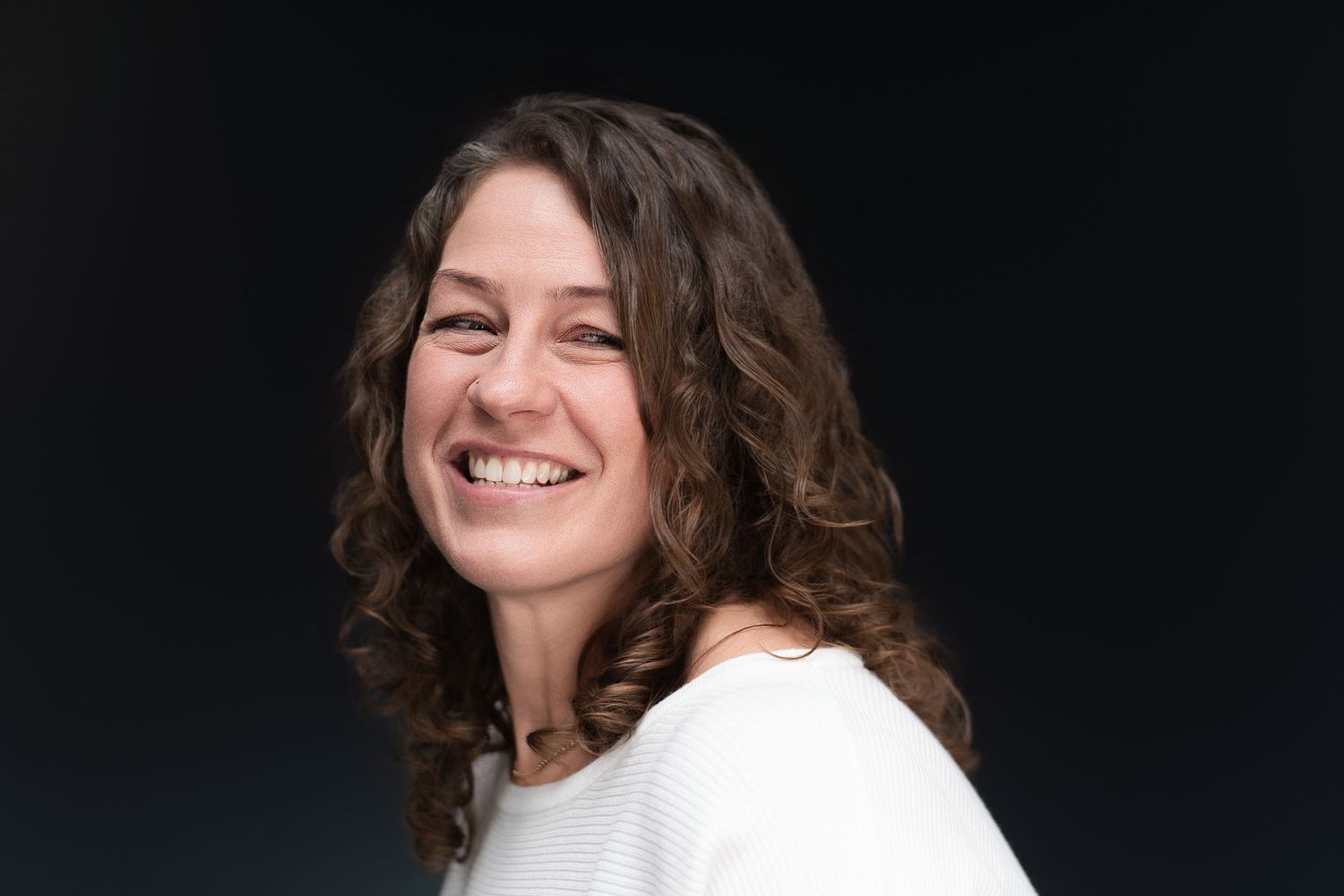 Side profile portrait of person with curly hair in white sweater smiling warmly.