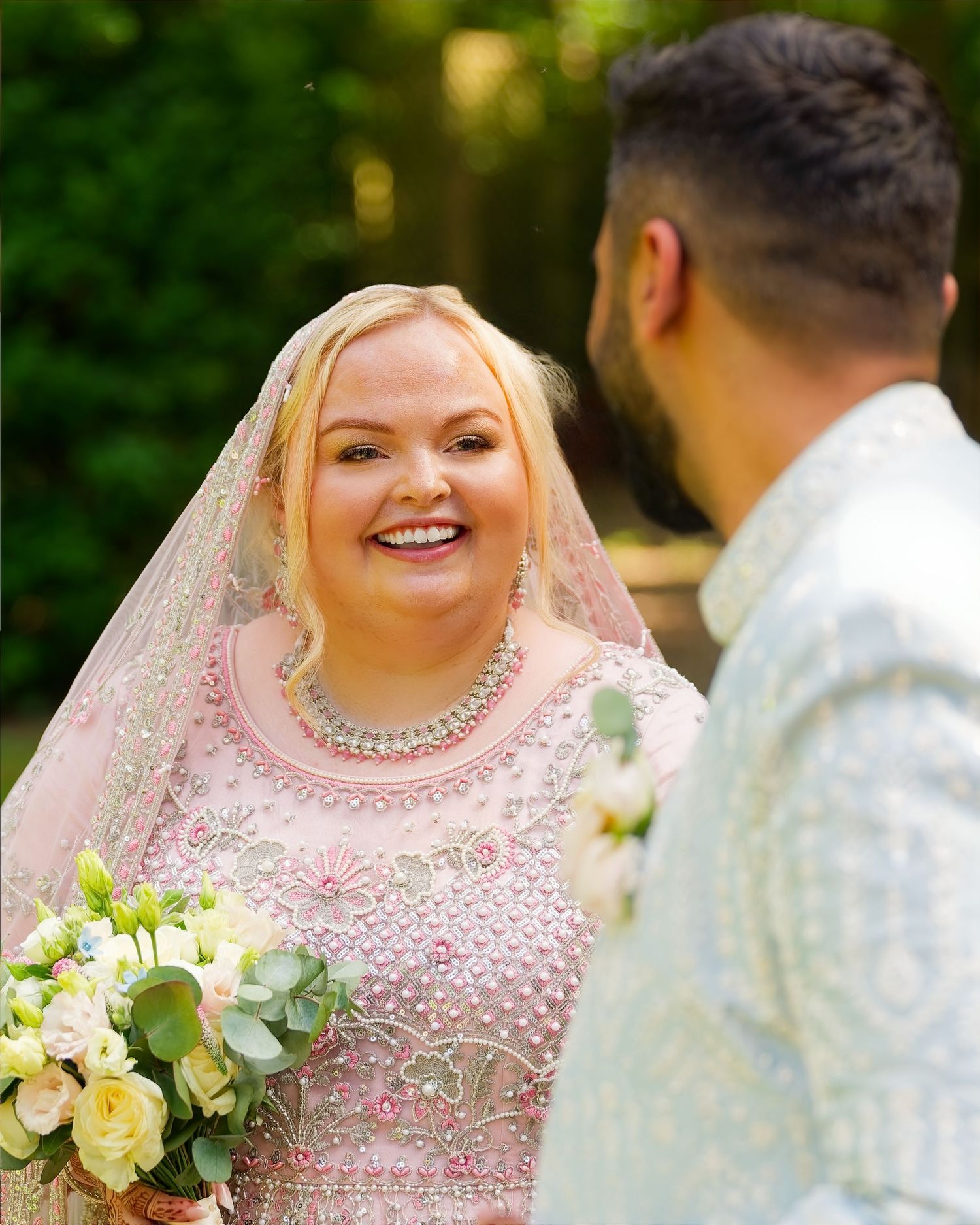 Smiling bride in pink embroidered lehenga with veil holds white bouquet facing groom outdoors.