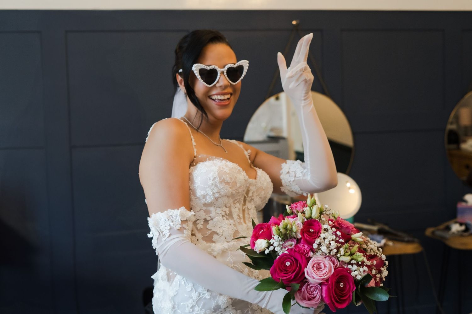 Joyful bride in white lace dress with heart sunglasses and gloves poses with bright pink floral bouquet.