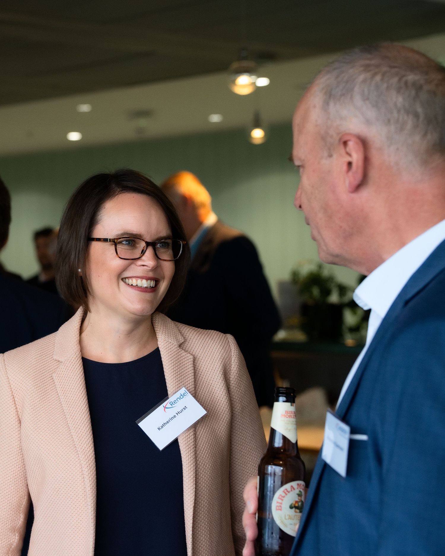 Smiling woman in a blazer and name tag chats with a man holding a beer bottle at a networking event.