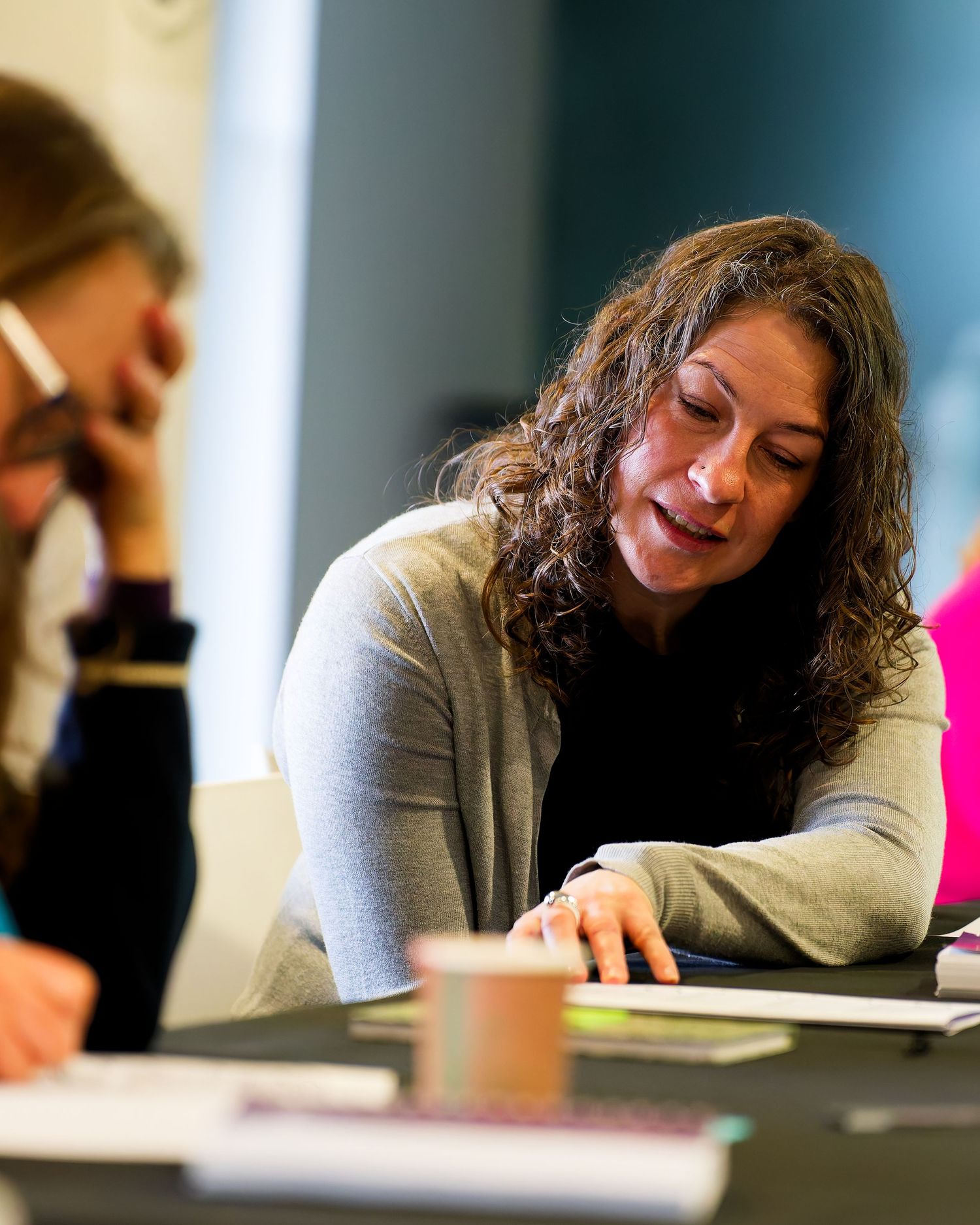 Smiling woman with curly brown hair in a gray cardigan looks down at papers at a conference table during a group meeting.