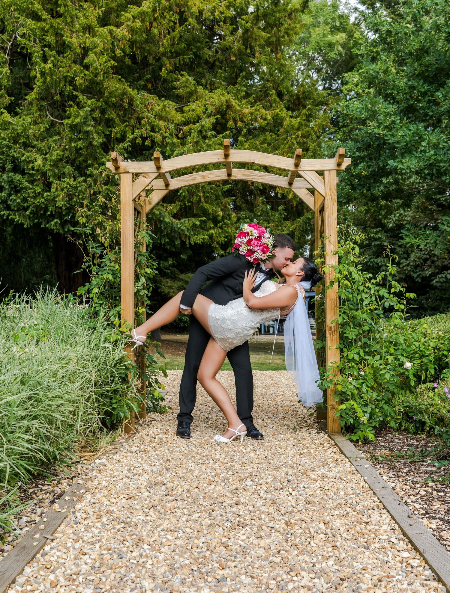 Groom dips bride for a romantic kiss under a wooden garden arch on a gravel path, bride holding pink bouquet.