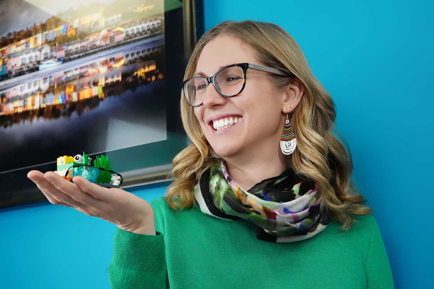Smiling woman in green sweater and glasses holds a small LEGO model, standing near a colorful framed photo on blue wall.