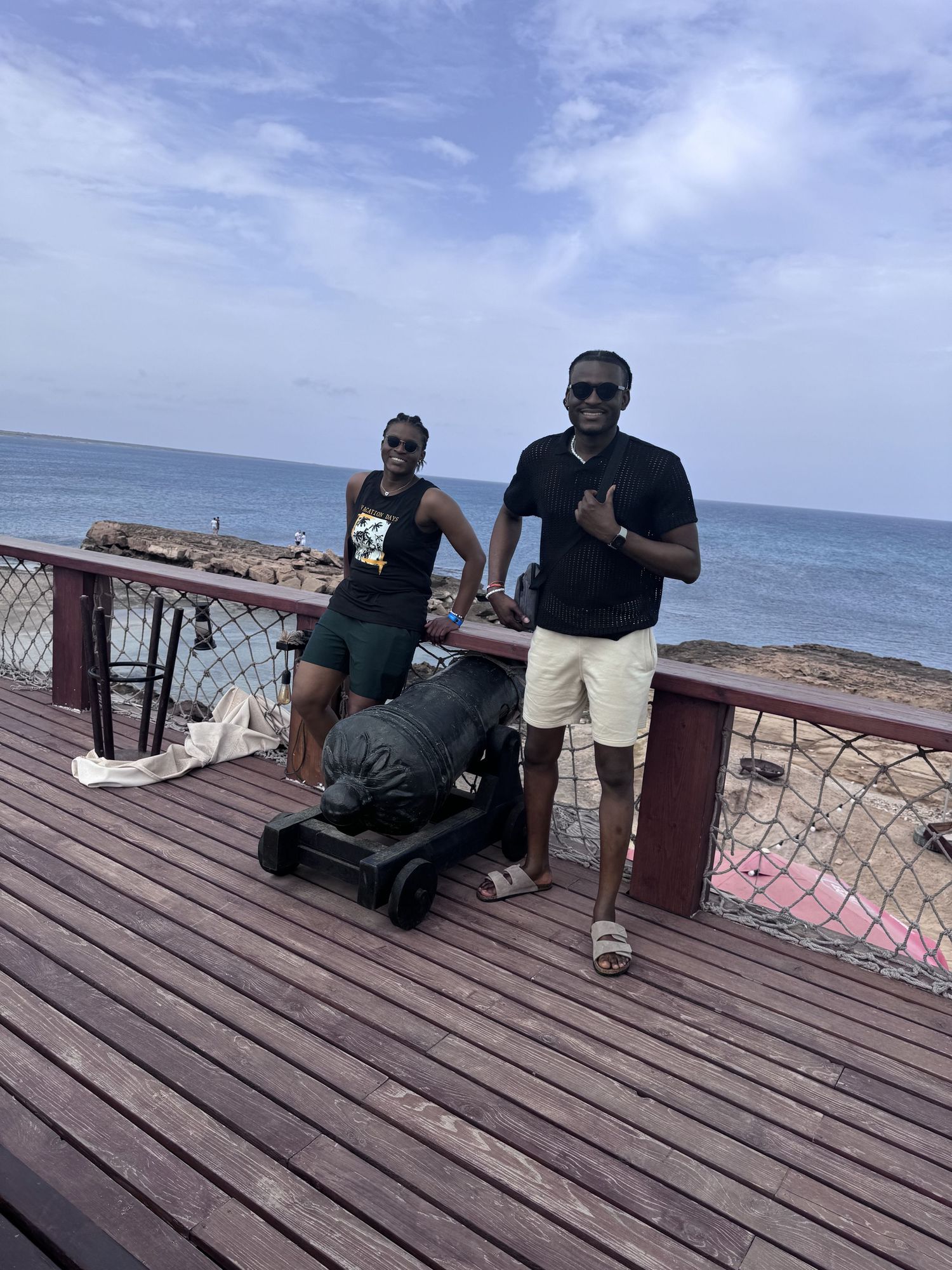 Two men in black shirts pose with an old cannon on a wooden deck overlooking the ocean.