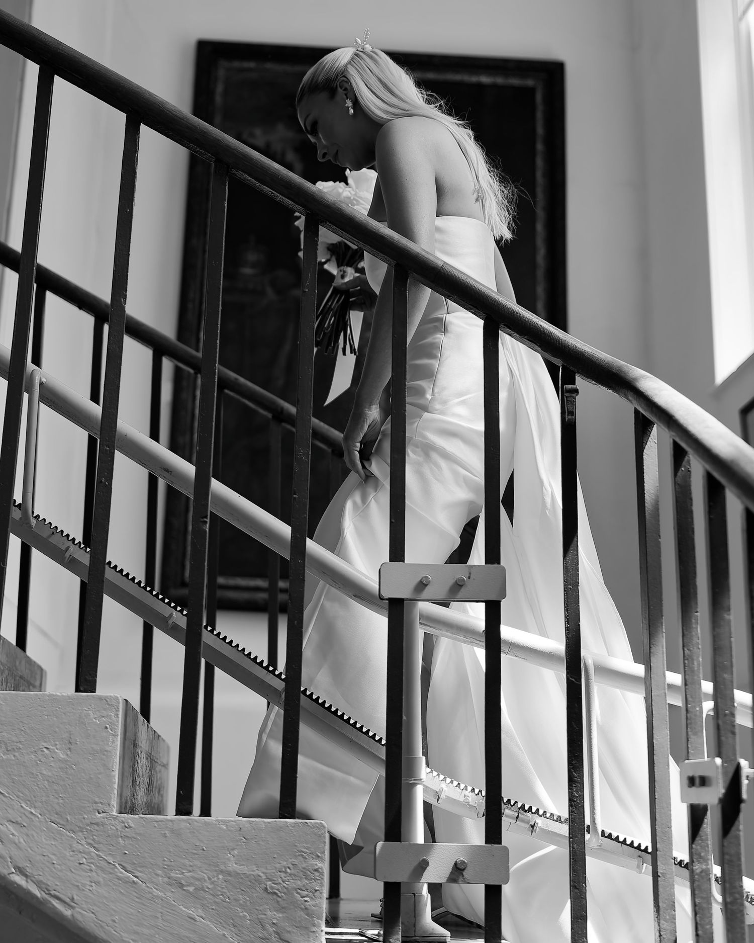 Black and white photo of a blonde bride in a white gown descending an ornate staircase with iron railings.
