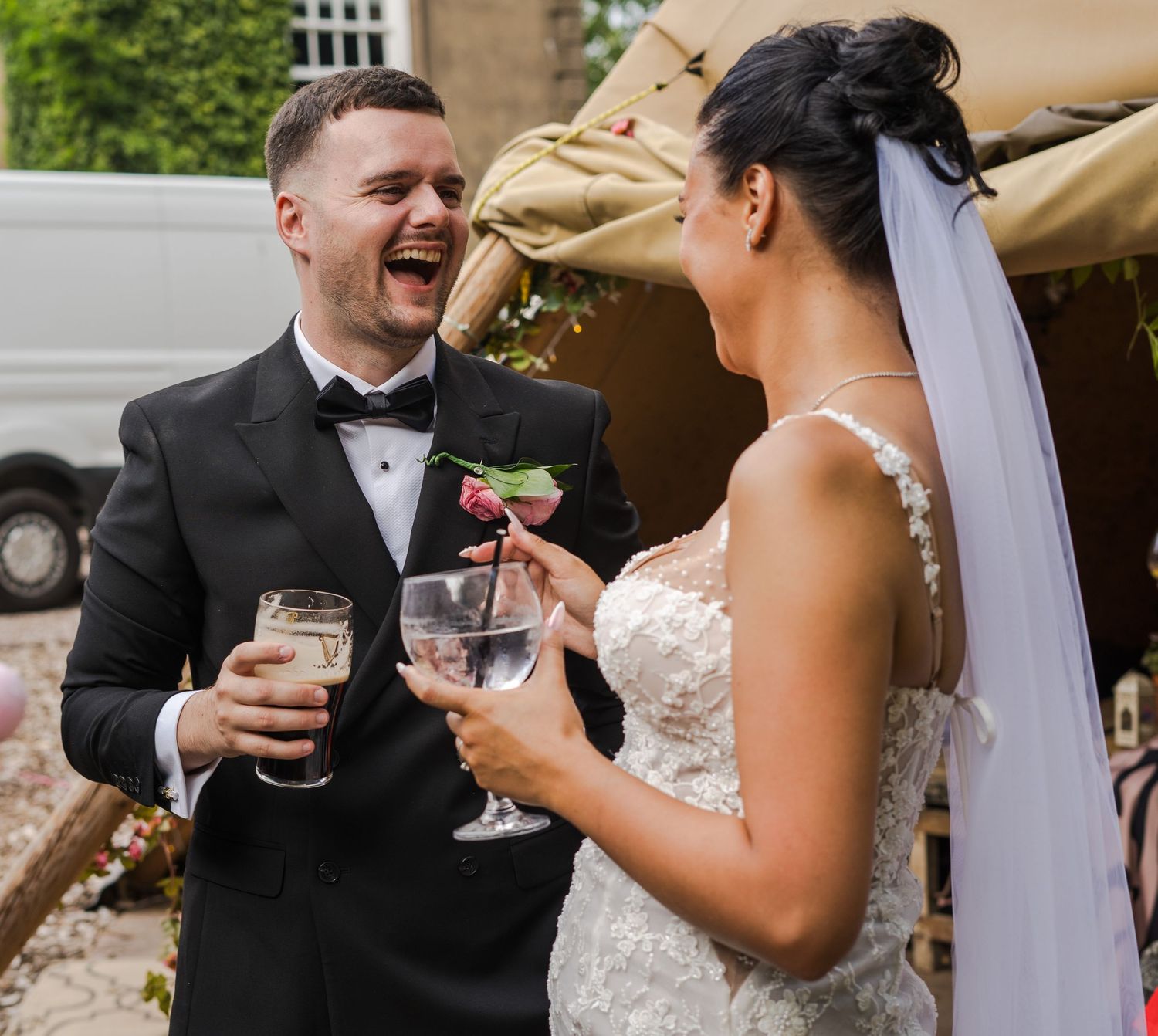 Laughing bride and groom in wedding attire toasting with cocktails outdoors at their wedding reception.