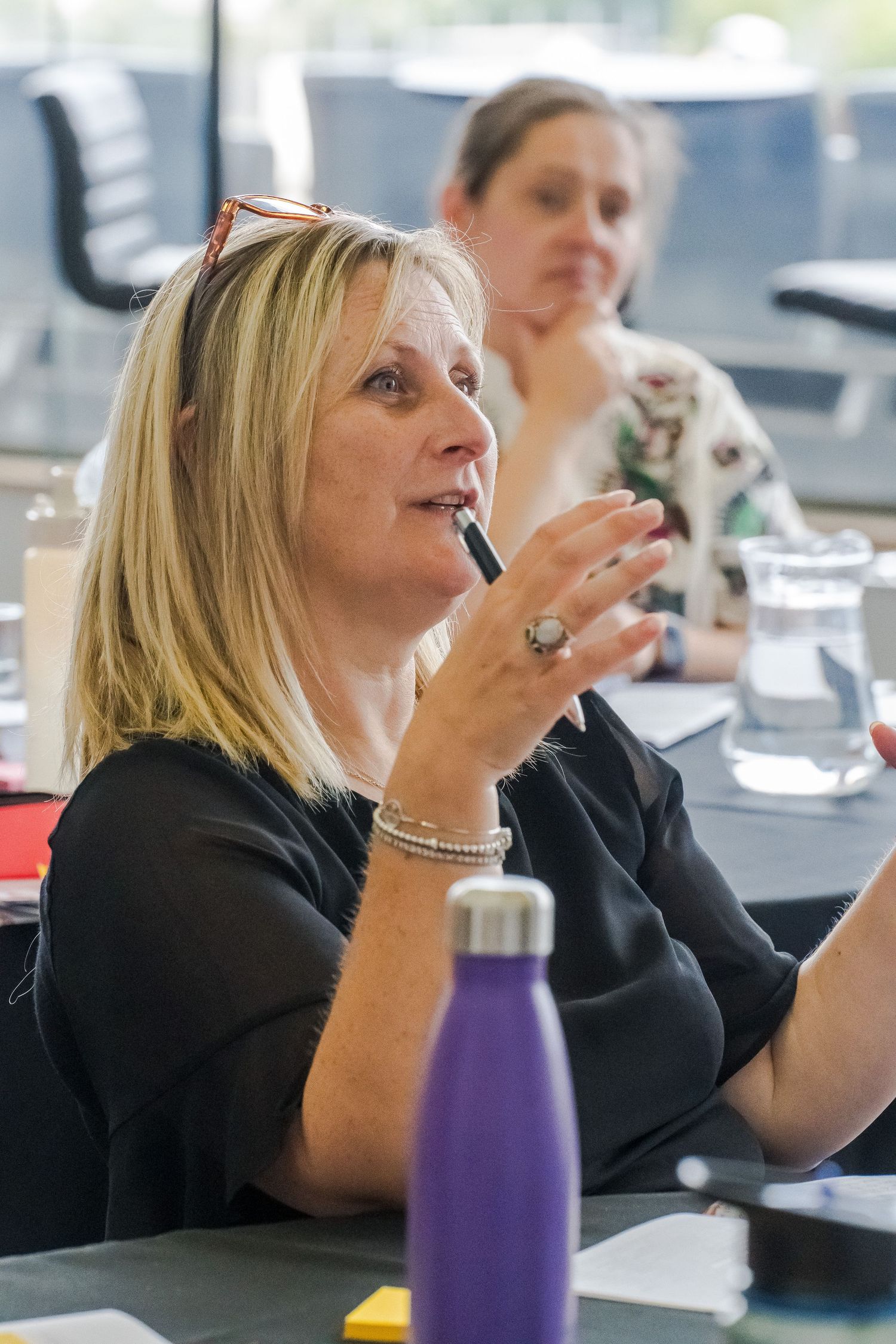 Blonde woman with sunglasses on her head gestures while speaking at a meeting table, with a purple water bottle and a colleague behind her.