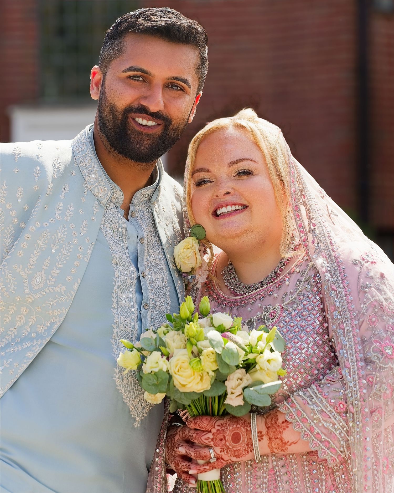 Couple in traditional South Asian wedding attire smiling, bride holding white bouquet with henna hands.
