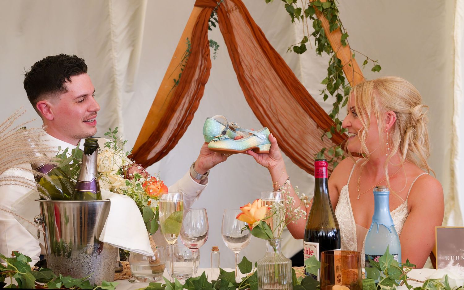 Bride and groom laughing together at wedding reception table decorated with flowers and greenery.