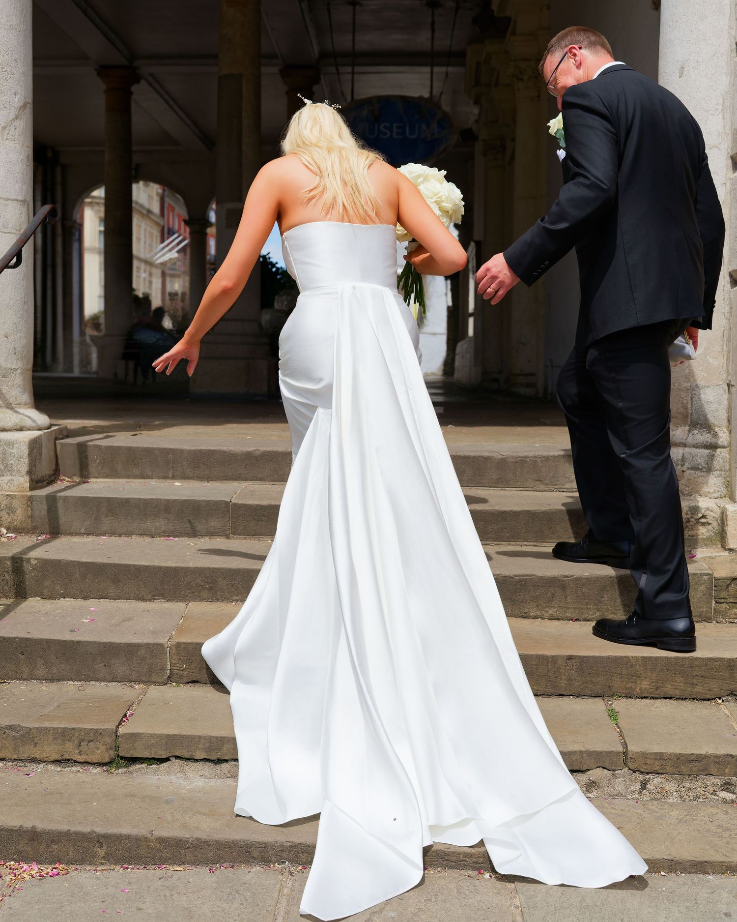 Bride in white strapless gown holding bouquet walks up stone steps with groom in black suit at wedding venue.