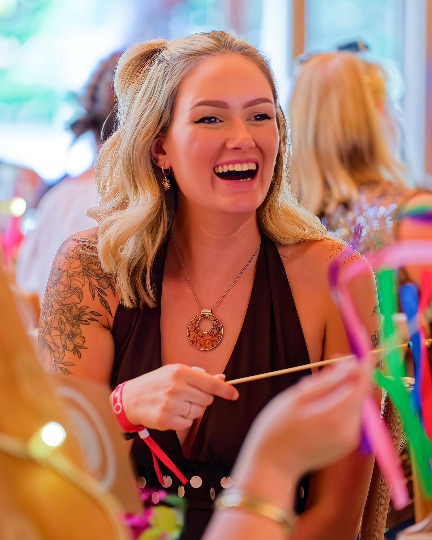 Smiling blonde woman with tattoos wearing a brown top and pendant necklace laughing at a colorful social event.