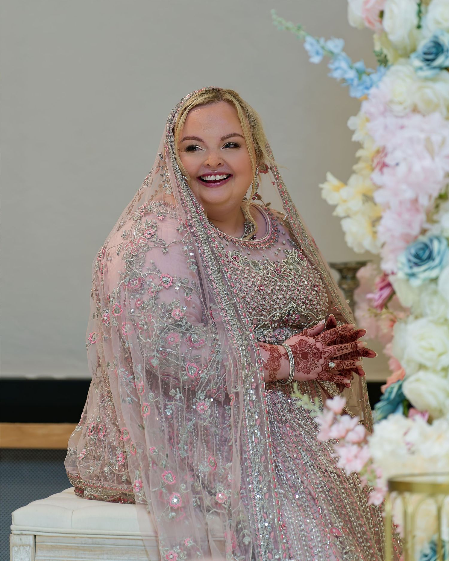 Smiling bride in pink embroidered lehenga with dupatta and mehndi, posing beside floral arrangement.