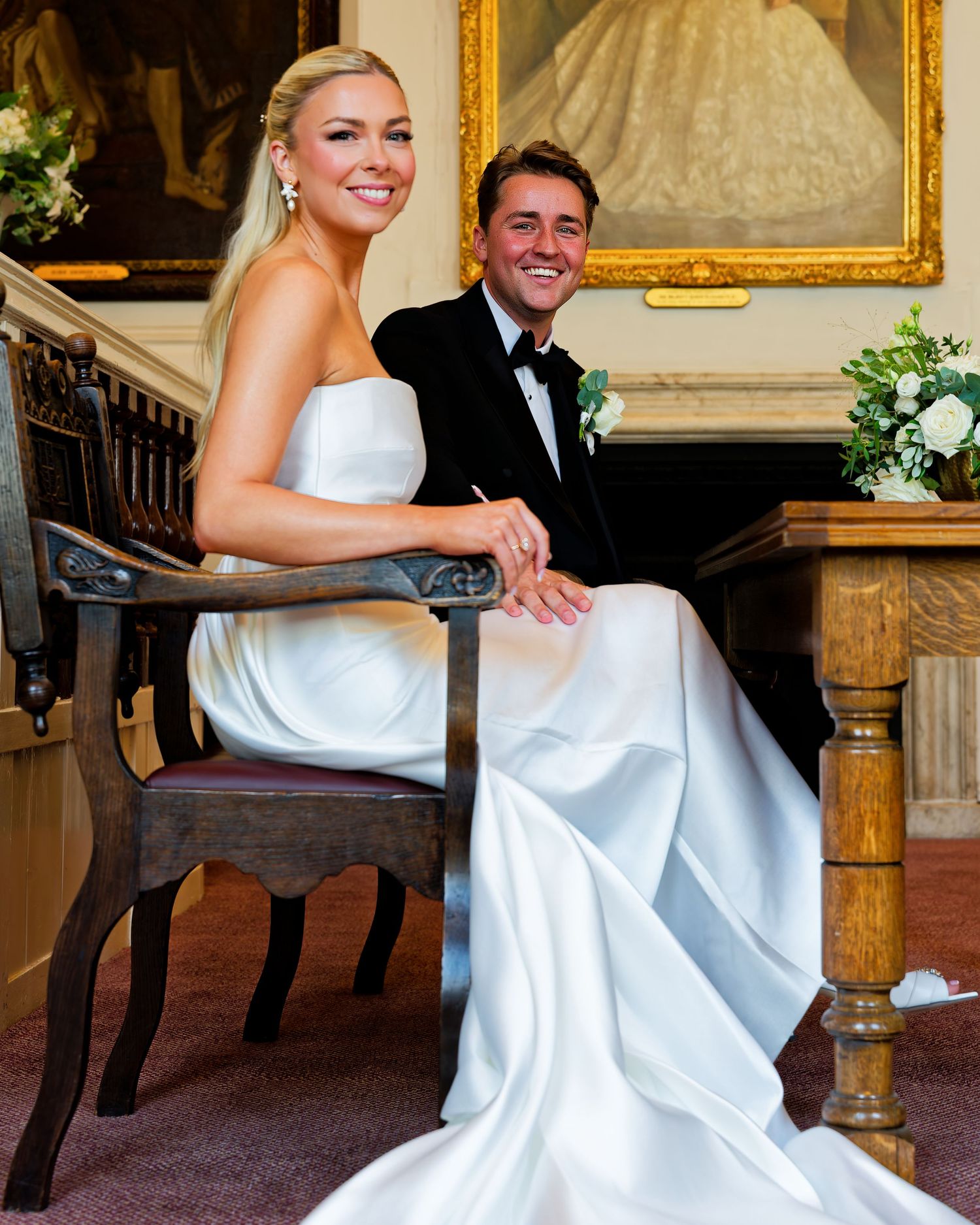 Elegant bride and groom in white satin gown and tuxedo seated at signing table.