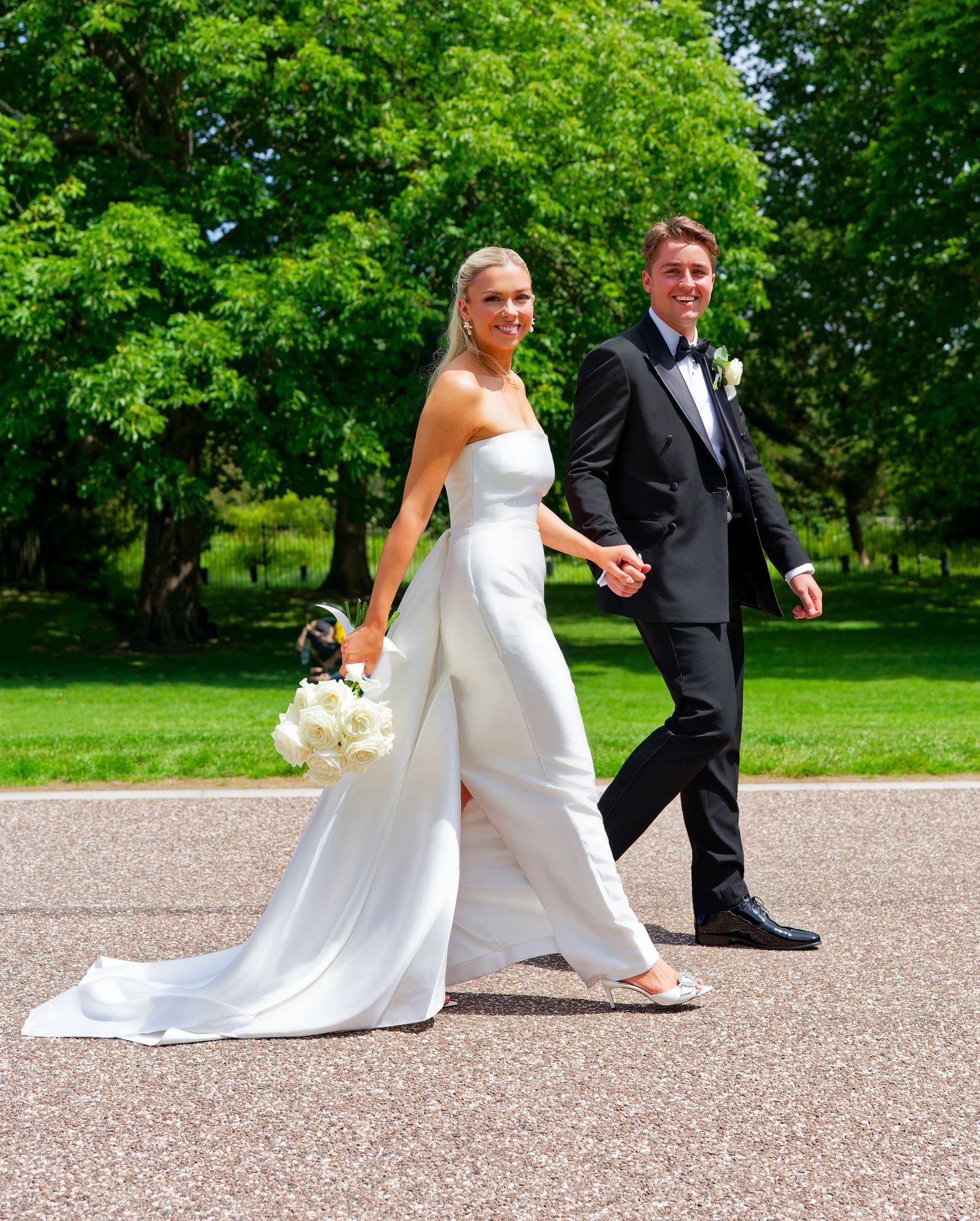 A bride in a white strapless gown holding a bouquet walks hand-in-hand with a groom in a black tuxedo outdoors.