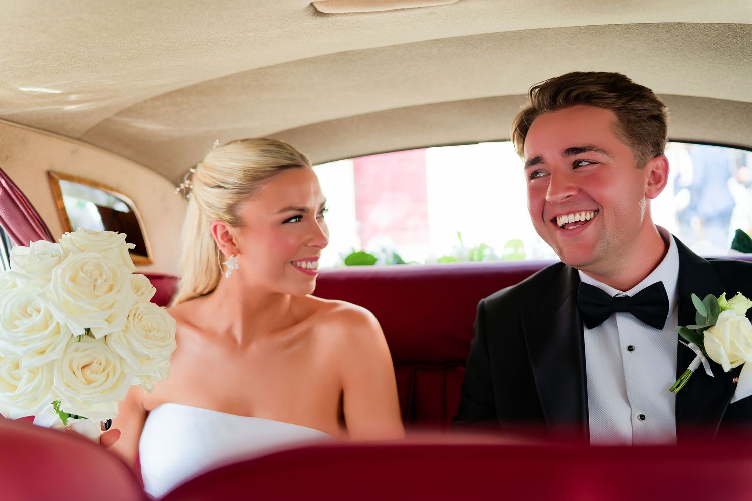 Happy bride and groom in wedding attire smiling at each other inside a classic car.