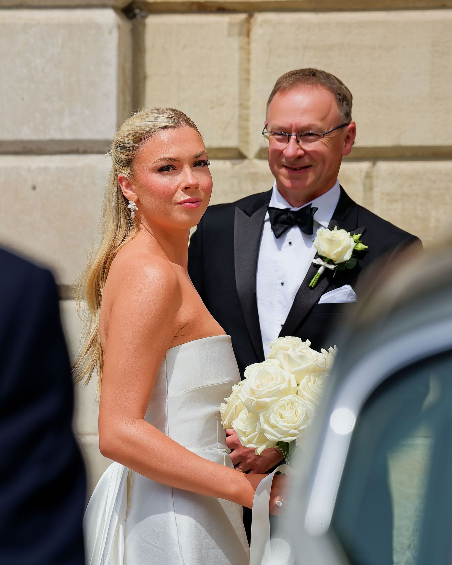 Bride in white strapless gown holding white roses bouquet stands with man in tuxedo at elegant wedding ceremony.