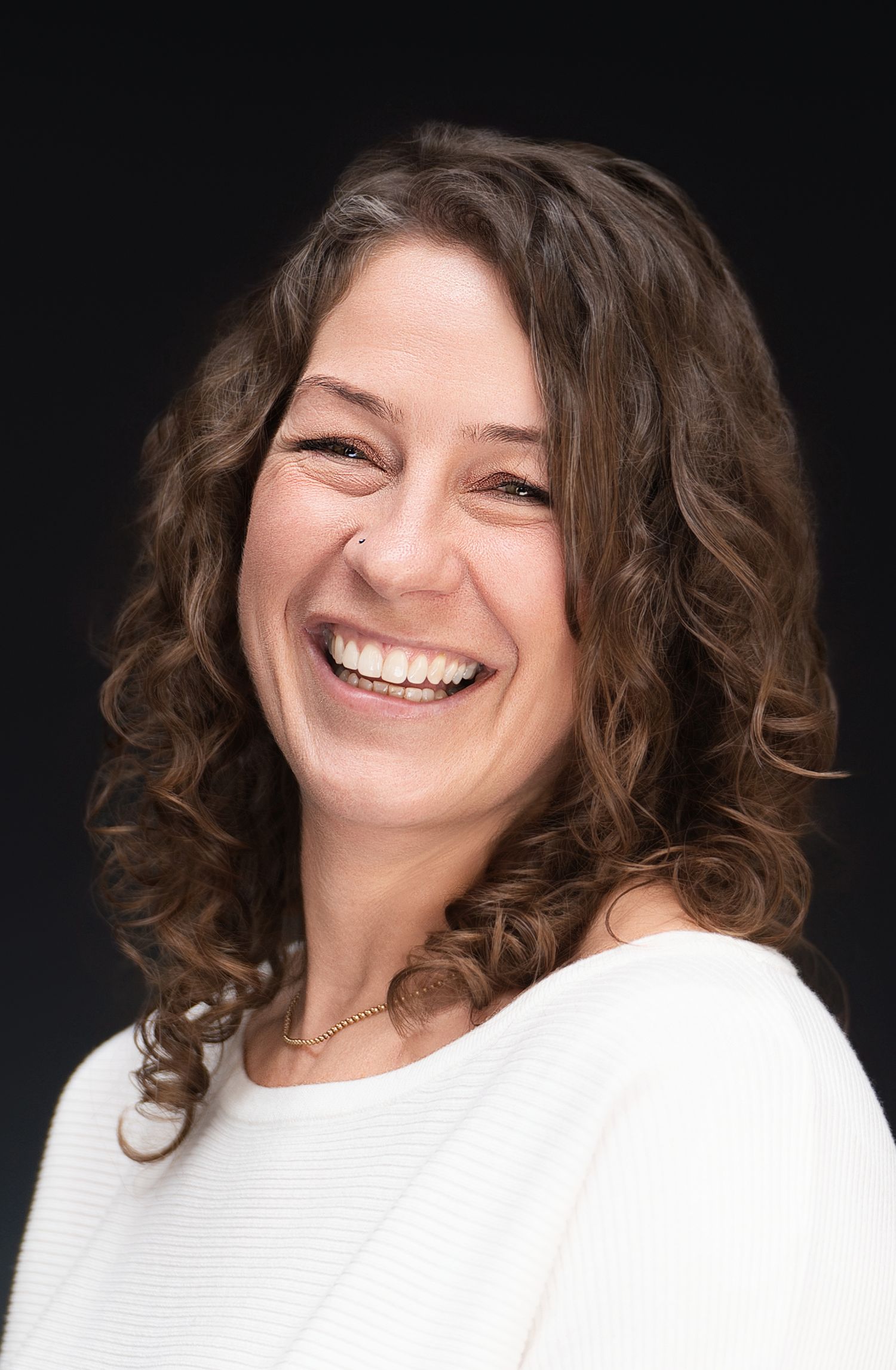 Portrait of person with curly hair wearing white sweater laughing joyfully against dark background.
