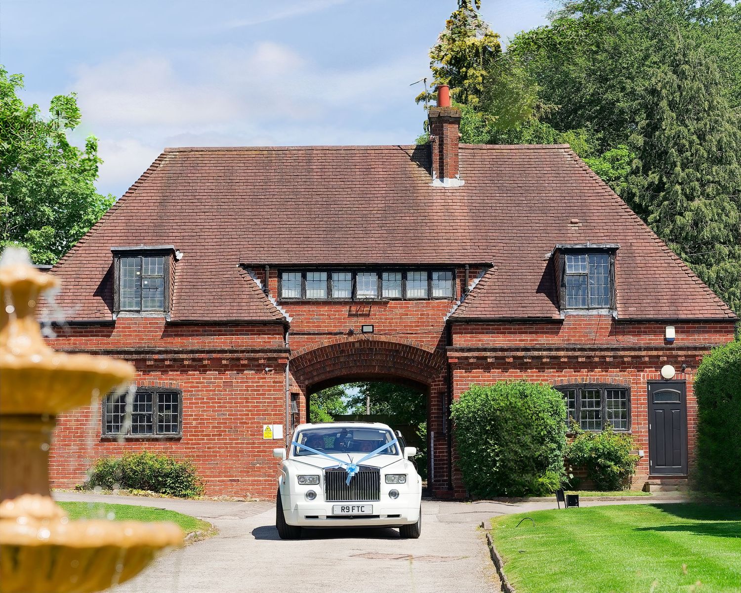 White Rolls-Royce wedding car parked under red brick archway entrance of elegant English manor venue.
