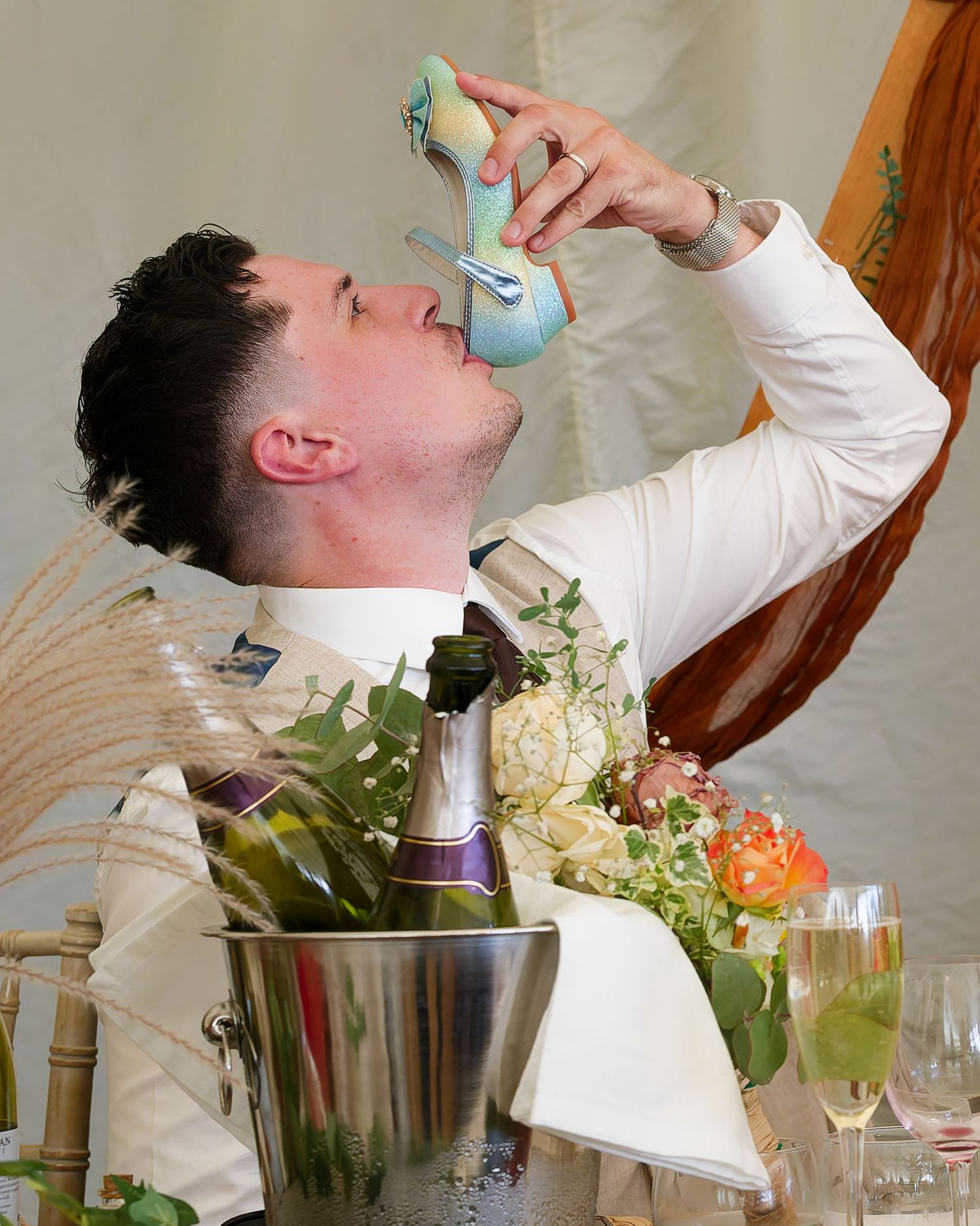 Man in white shirt drinking champagne from a high heel shoe at a wedding reception with flowers and bubbly.