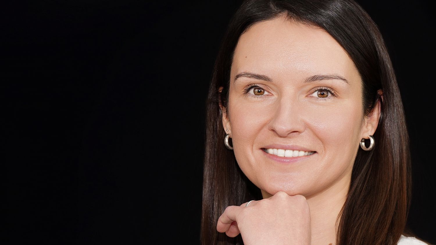 Professional headshot of person with dark hair smiling warmly against black background.