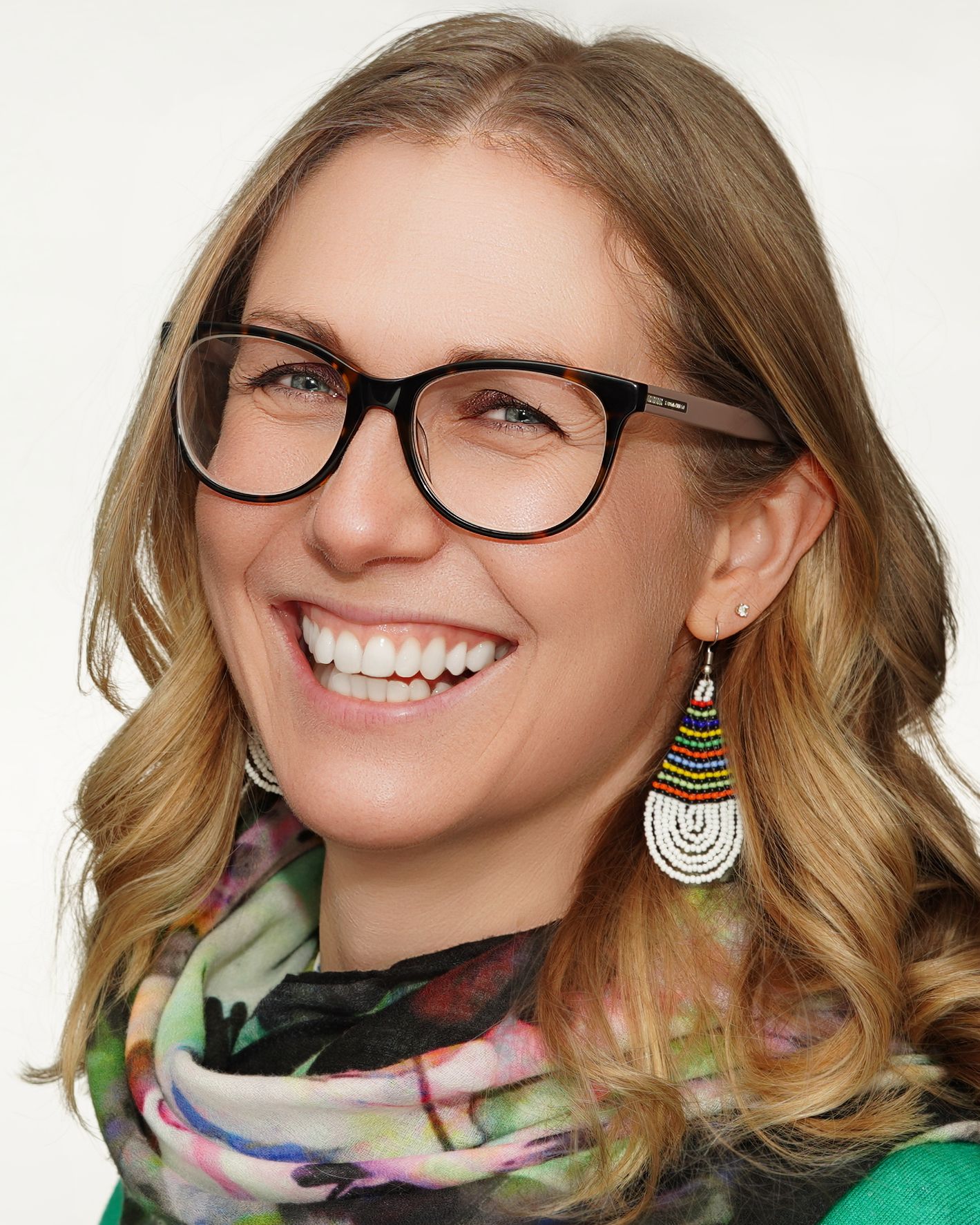 A smiling person wearing black-framed glasses and colorful layered earrings with a patterned scarf around their neck.