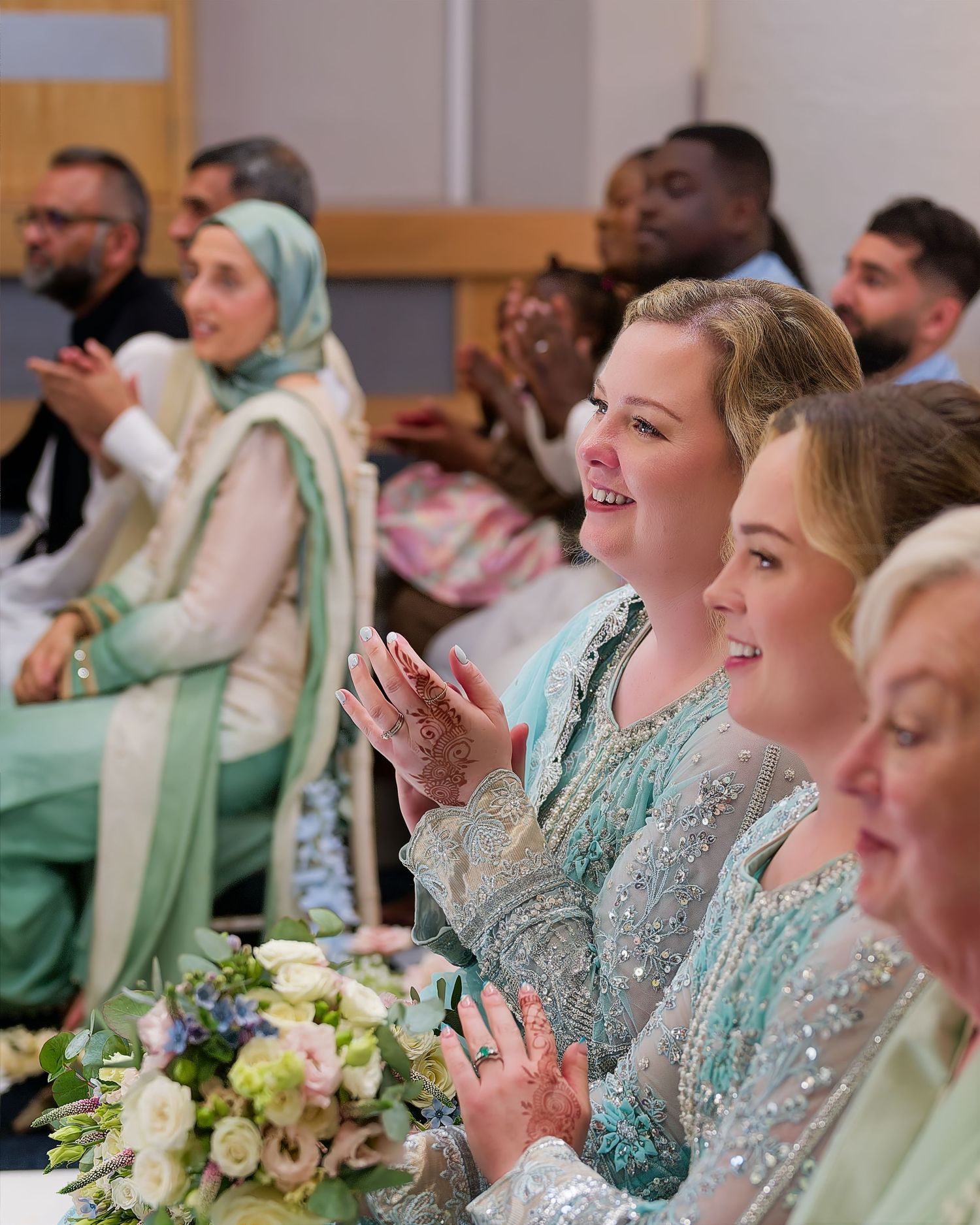 Wedding guests in elegant outfits clapping and smiling during a joyful South Asian wedding celebration.