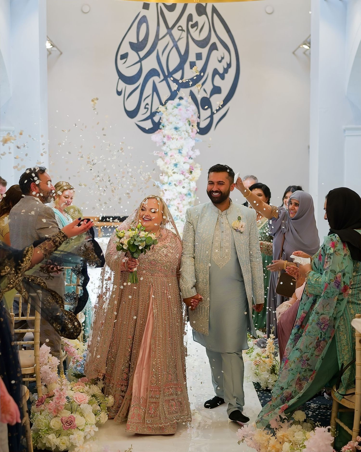 Couple walks down aisle at South Asian wedding ceremony with confetti, floral decor, and Arabic calligraphy backdrop.