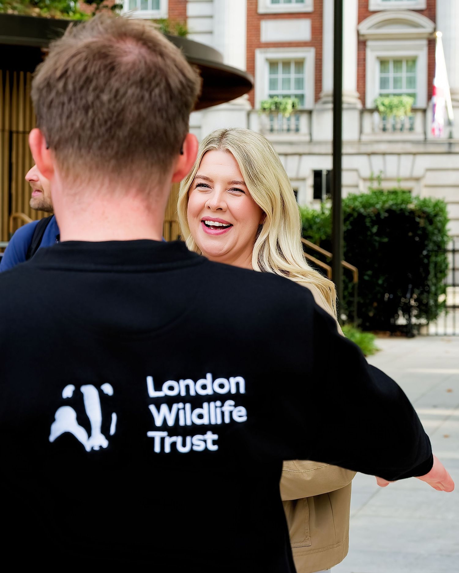 Smiling woman with blonde hair greets a person wearing a black London Wildlife Trust t-shirt on a city street near brick buildings.