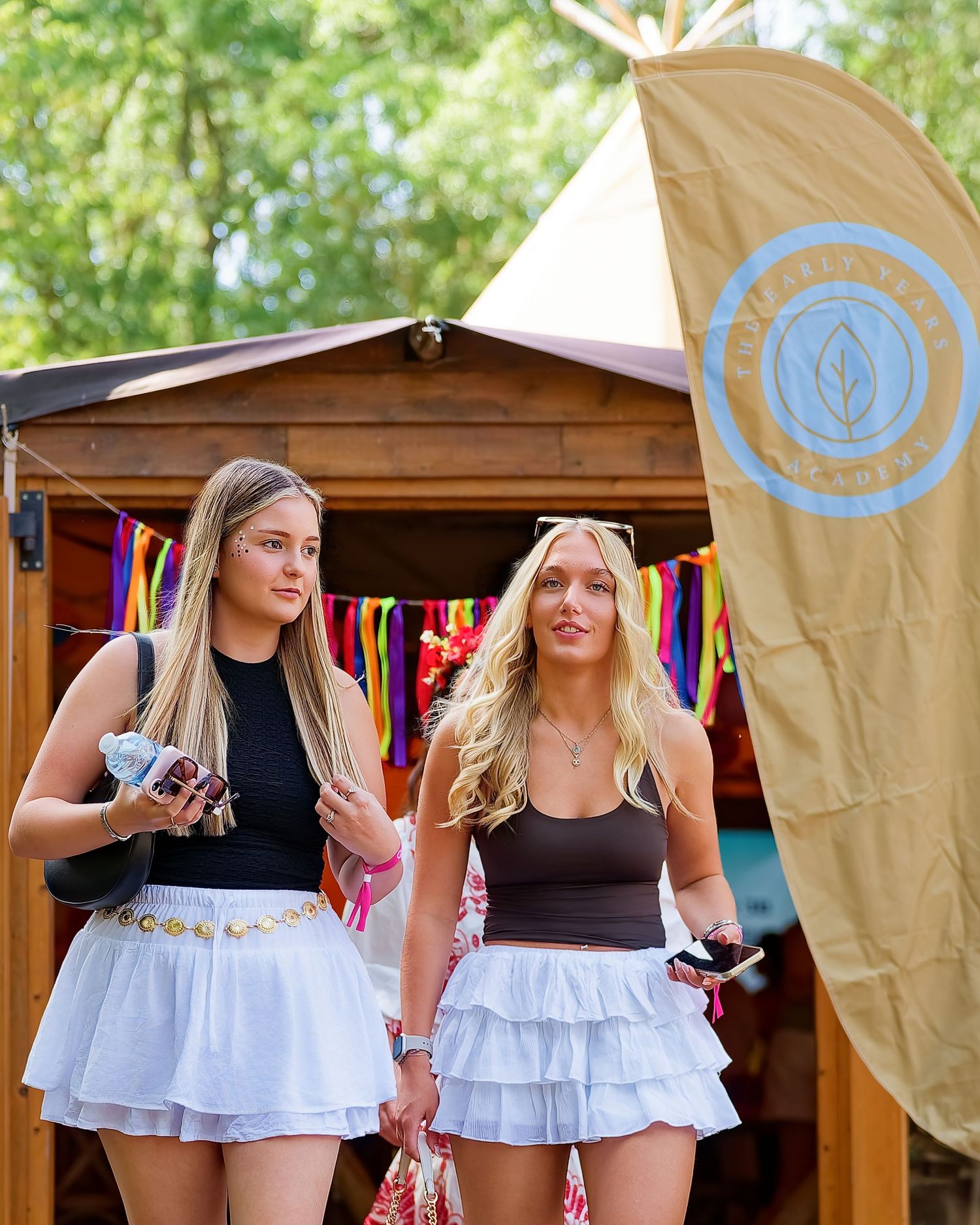 Two young women in white ruffled skirts and crop tops walk past a wooden hut with colorful bunting at an outdoor festival.
