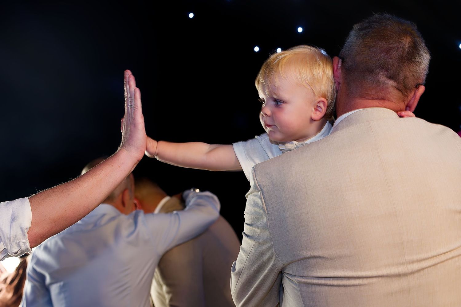 Blonde toddler boy in beige suit giving a high five at a wedding celebration event.