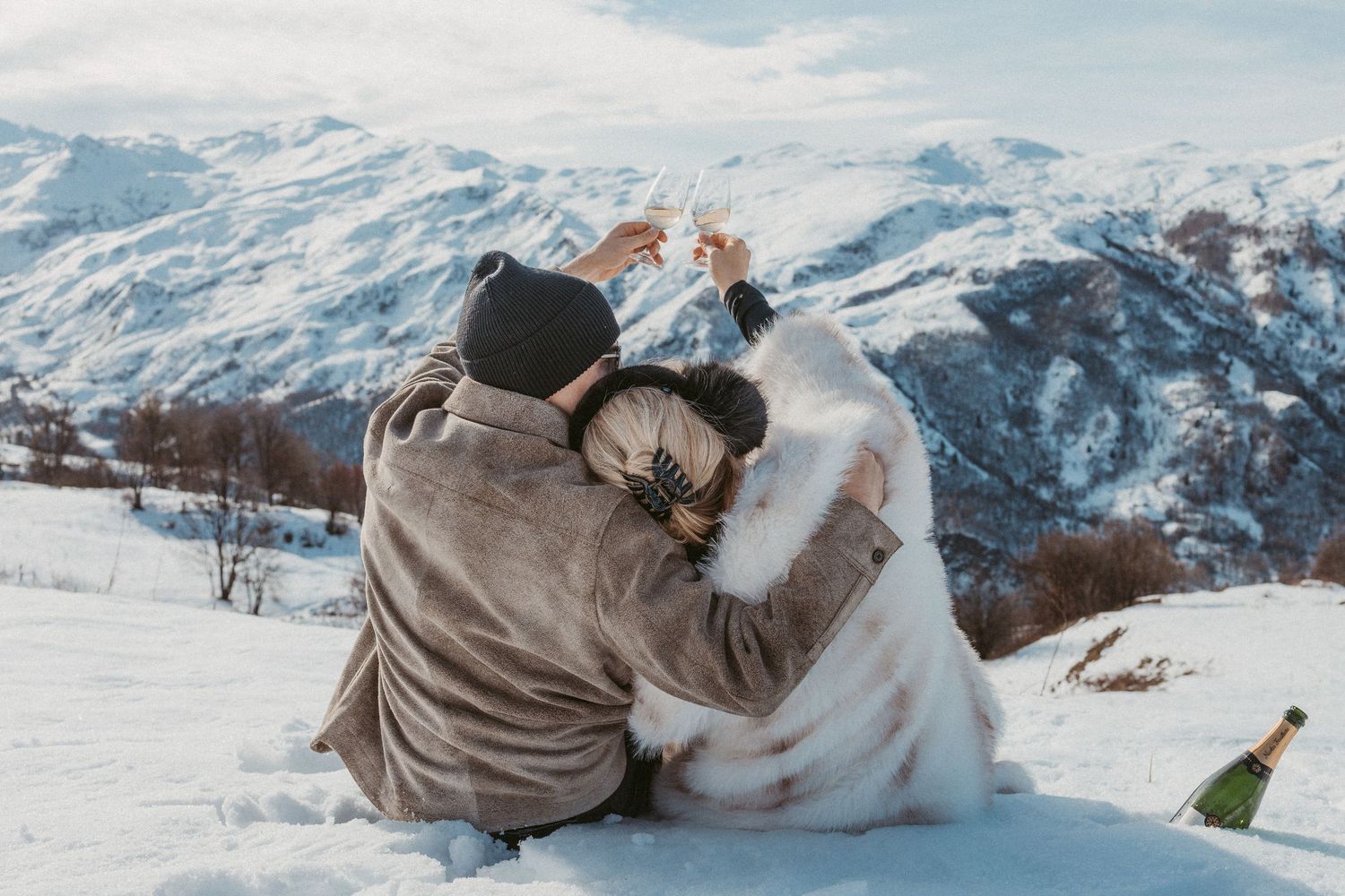 newly engaged couple sit in snow toasting champagne glasses above their heads while looking at mountain views