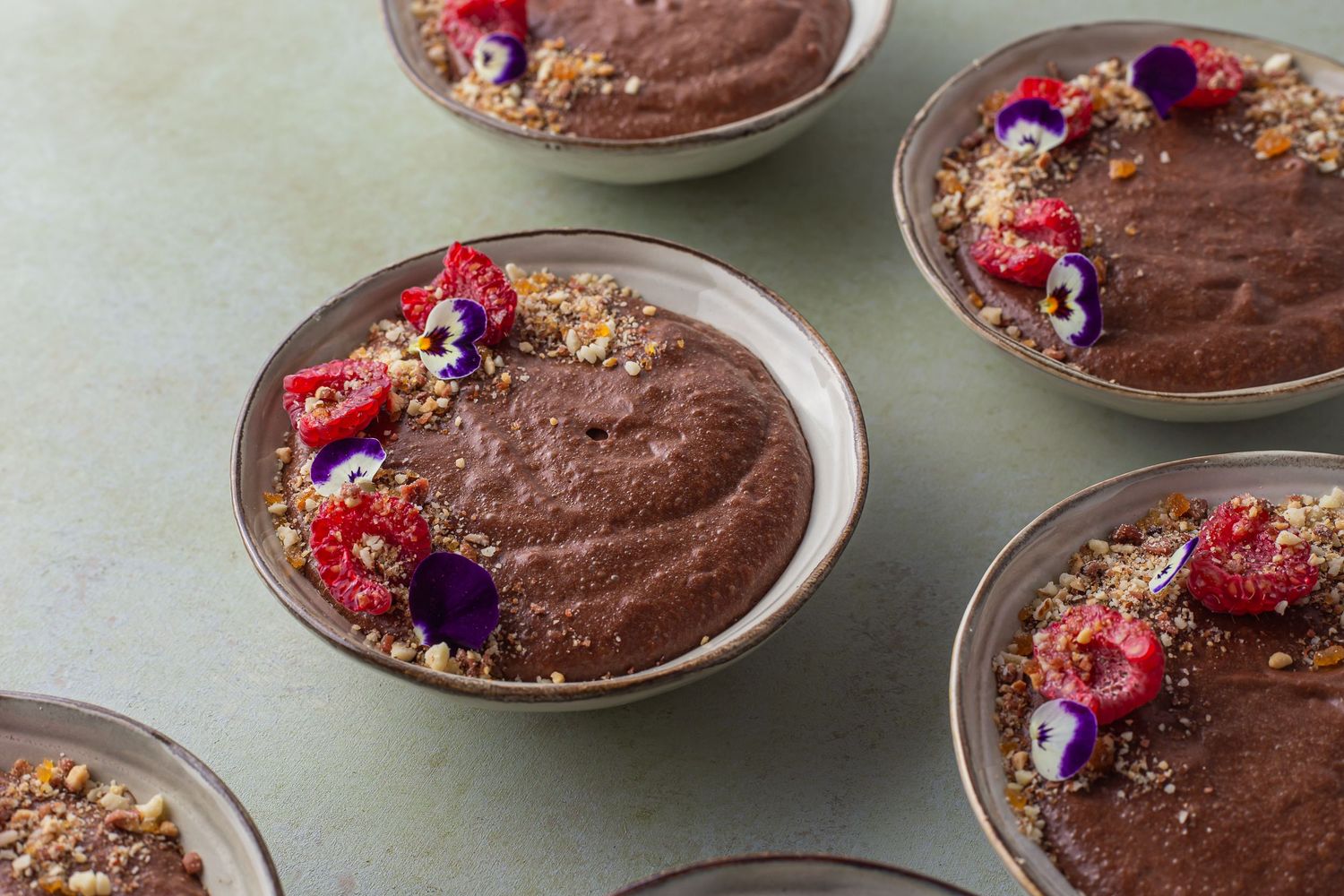 multiple chocolate mousses served in individual bowls, garnished with hazel nut crumb , raspberries and edible flowers
