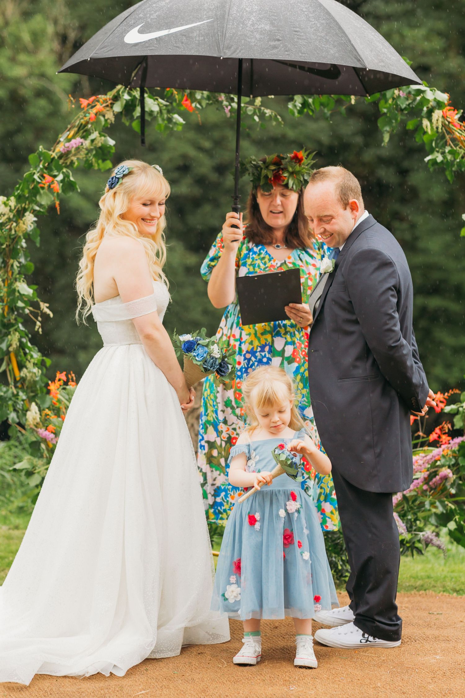 bride and groom look at daughter stood between them at altar while officiant holds large black umbrella