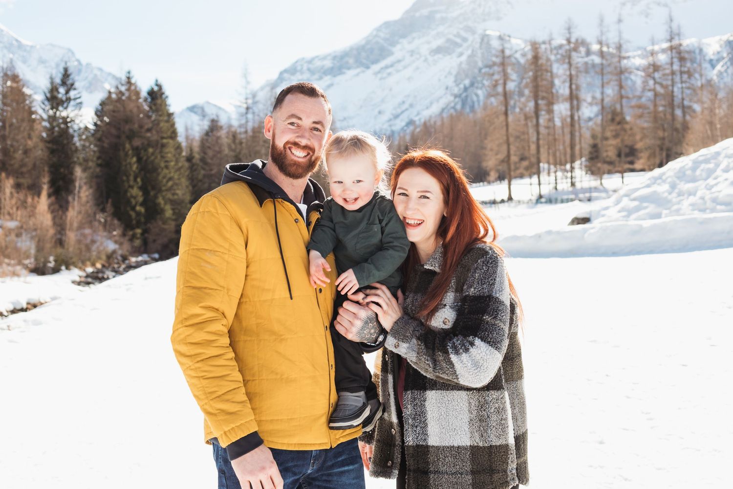 pregnant mother and husband snuggle their toddler son outside in the snow