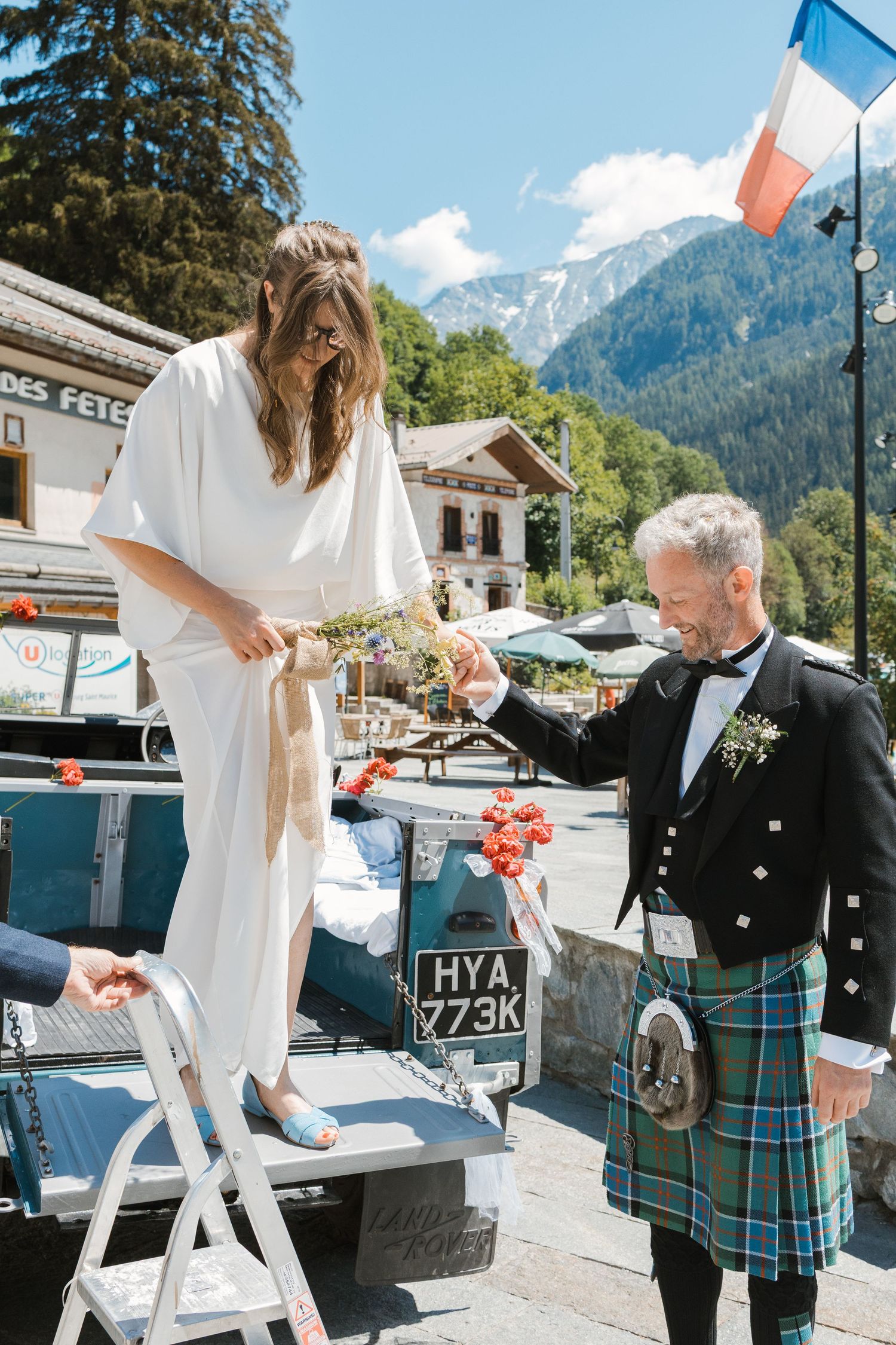 Scottish groom wearing kilt helps bride step down from open top land rover
