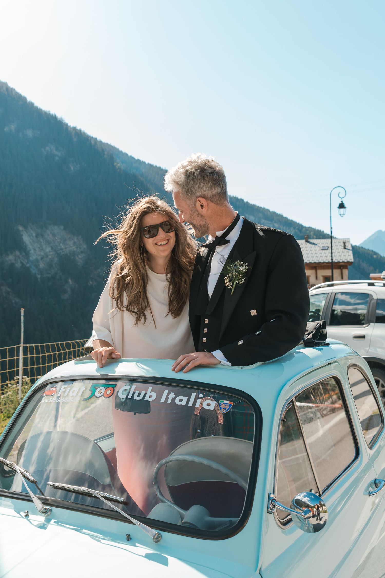 bride and groom stands through sun roof of vintage blue fiat car