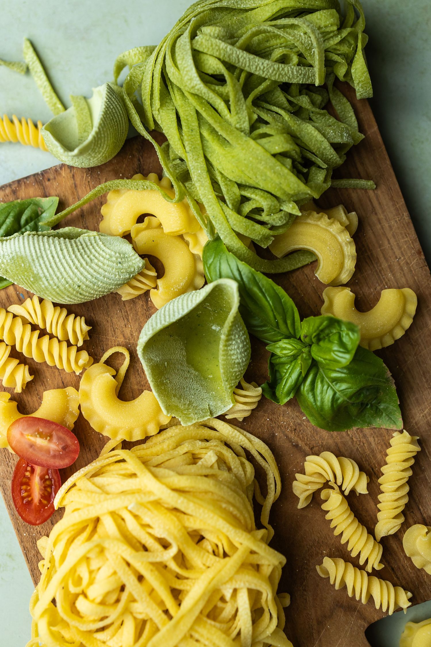 wooden chopping board with fresh green and traditional coloured pasta and basil and tomatoes