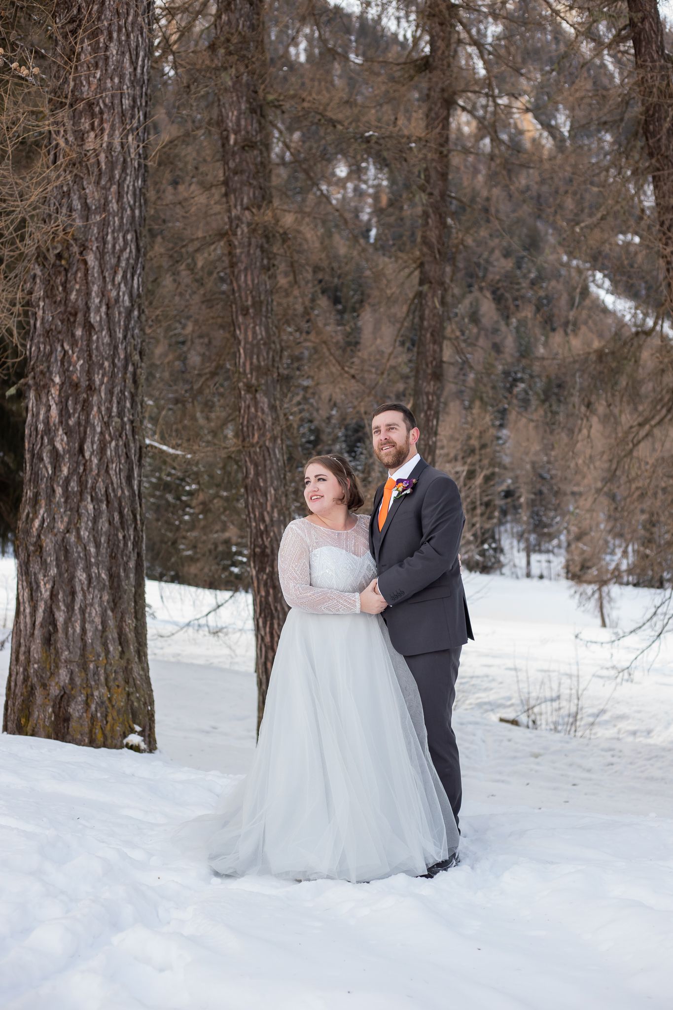 bride and groom stand in snow surrounded by trees looking to the mountains